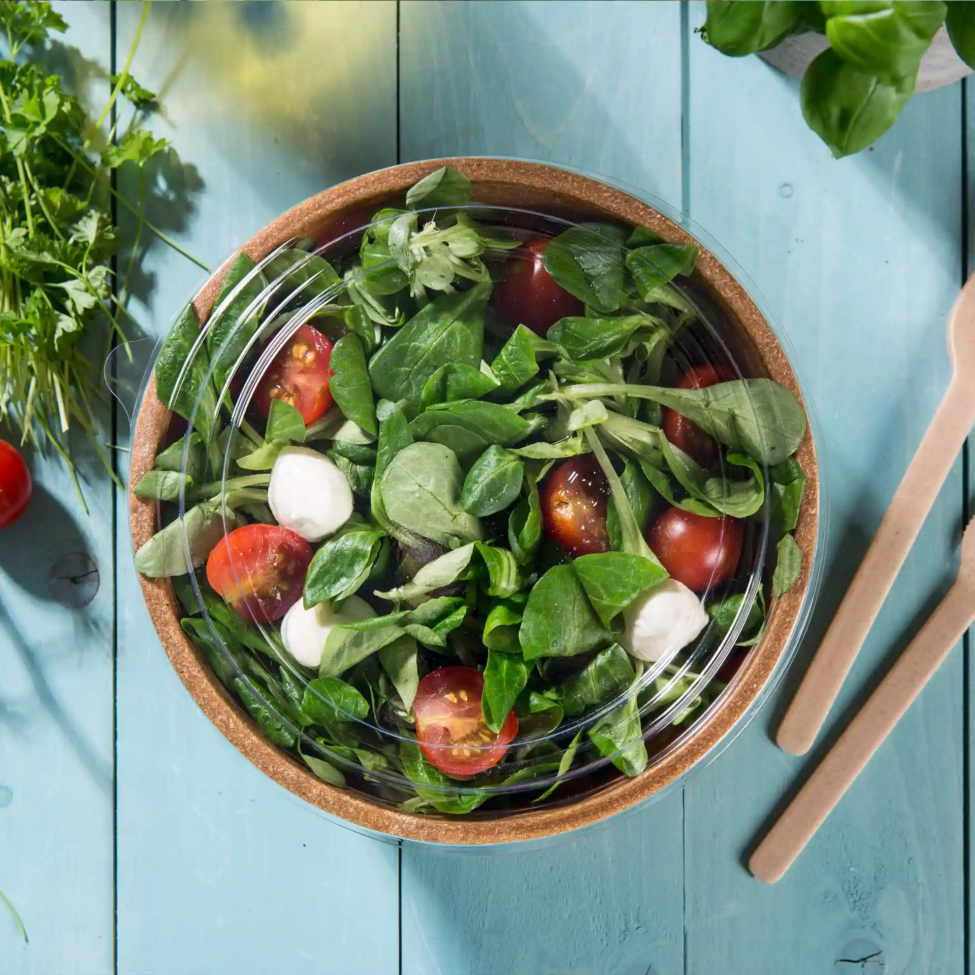 Klarer runder Becherdeckel aus PLA, auf einer Schüssel mit Salat, Tomaten und Spinat auf einer blauen Holzoberfläche.
