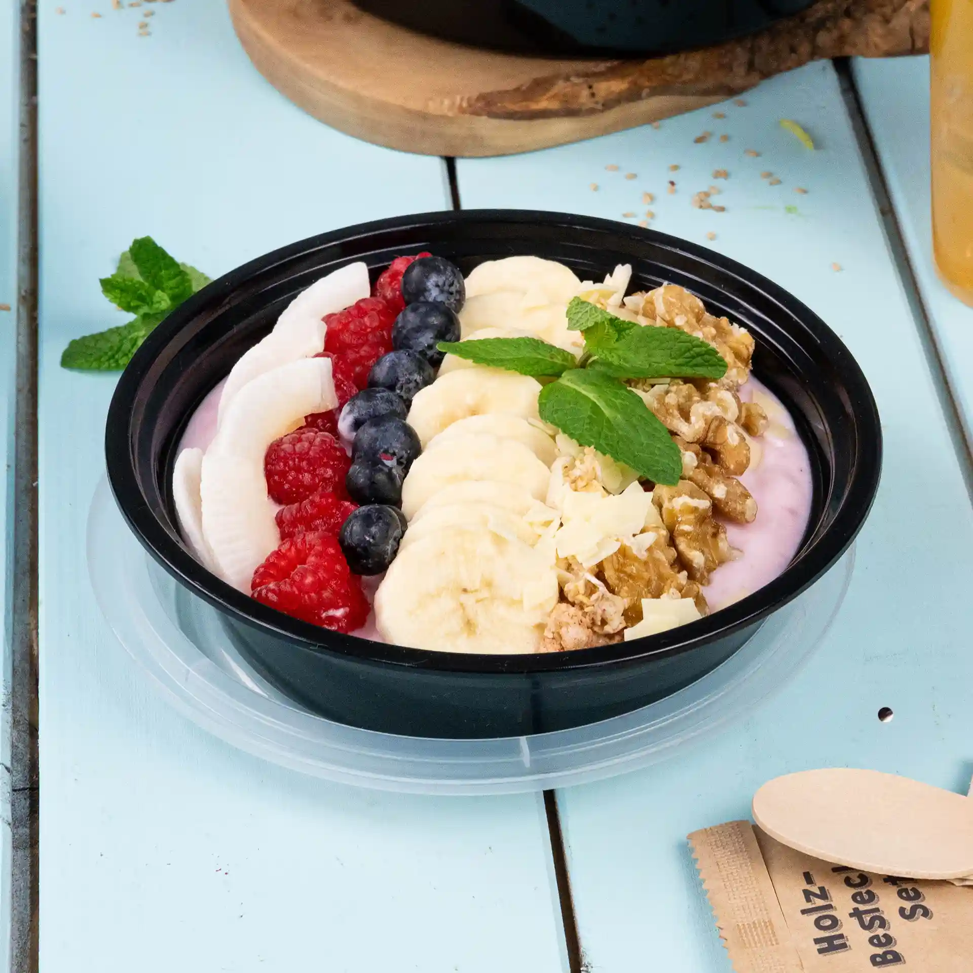 Schüssel gefüllt mit Joghurt, Bananenscheiben, Himbeeren, Heidelbeeren, Granola und Minzblättern auf einer hellblauen Oberfläche.
