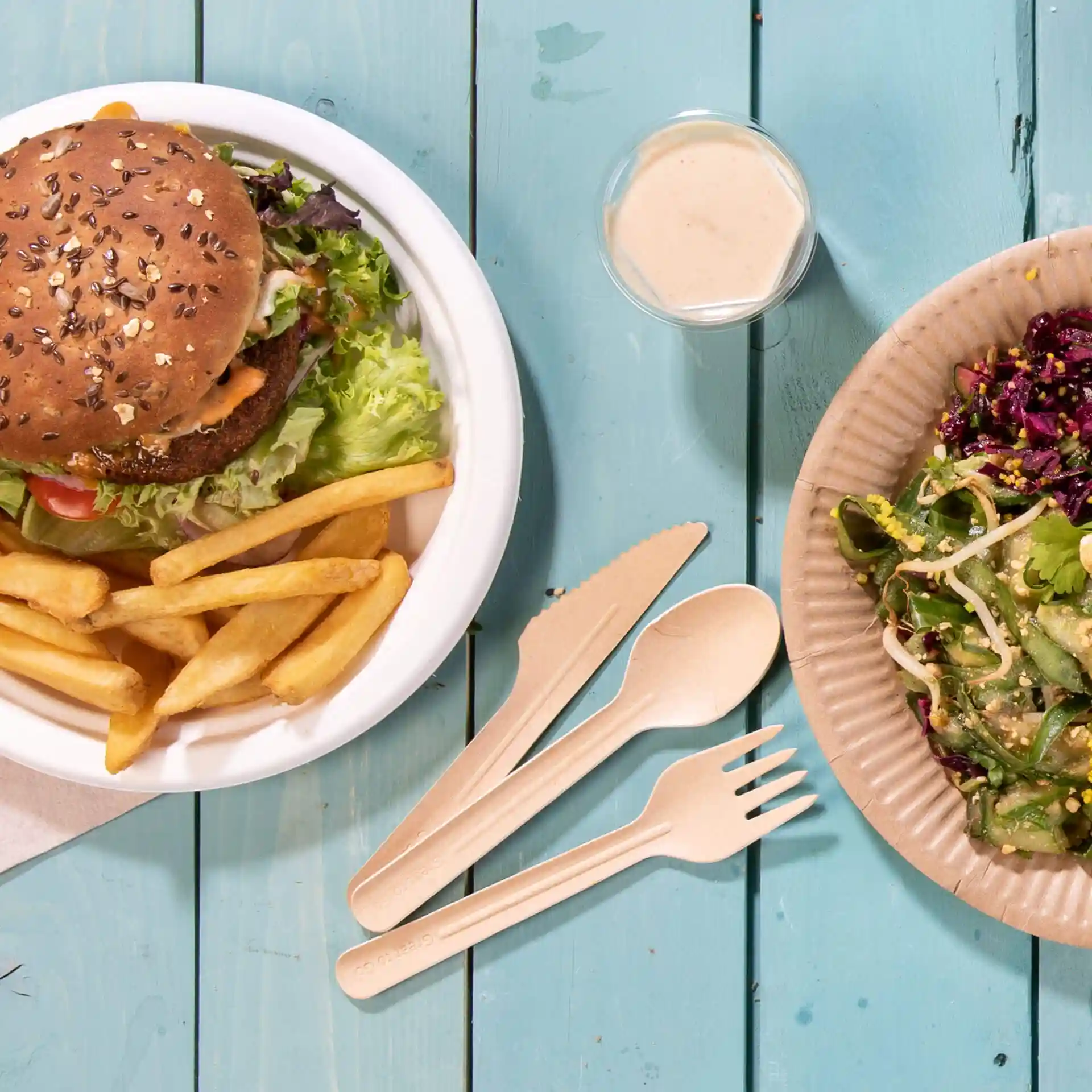 Bild eines Burgers mit Salat und Pommes in einer weißen Schüssel, dazu ein Salat auf einem braunen Pappteller und Holzbesteck.