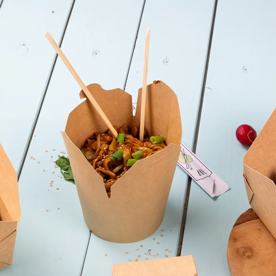 Takeout box filled with noodles, topped with green peppers and sesame seeds, with chopsticks inserted, on a light blue surface.
