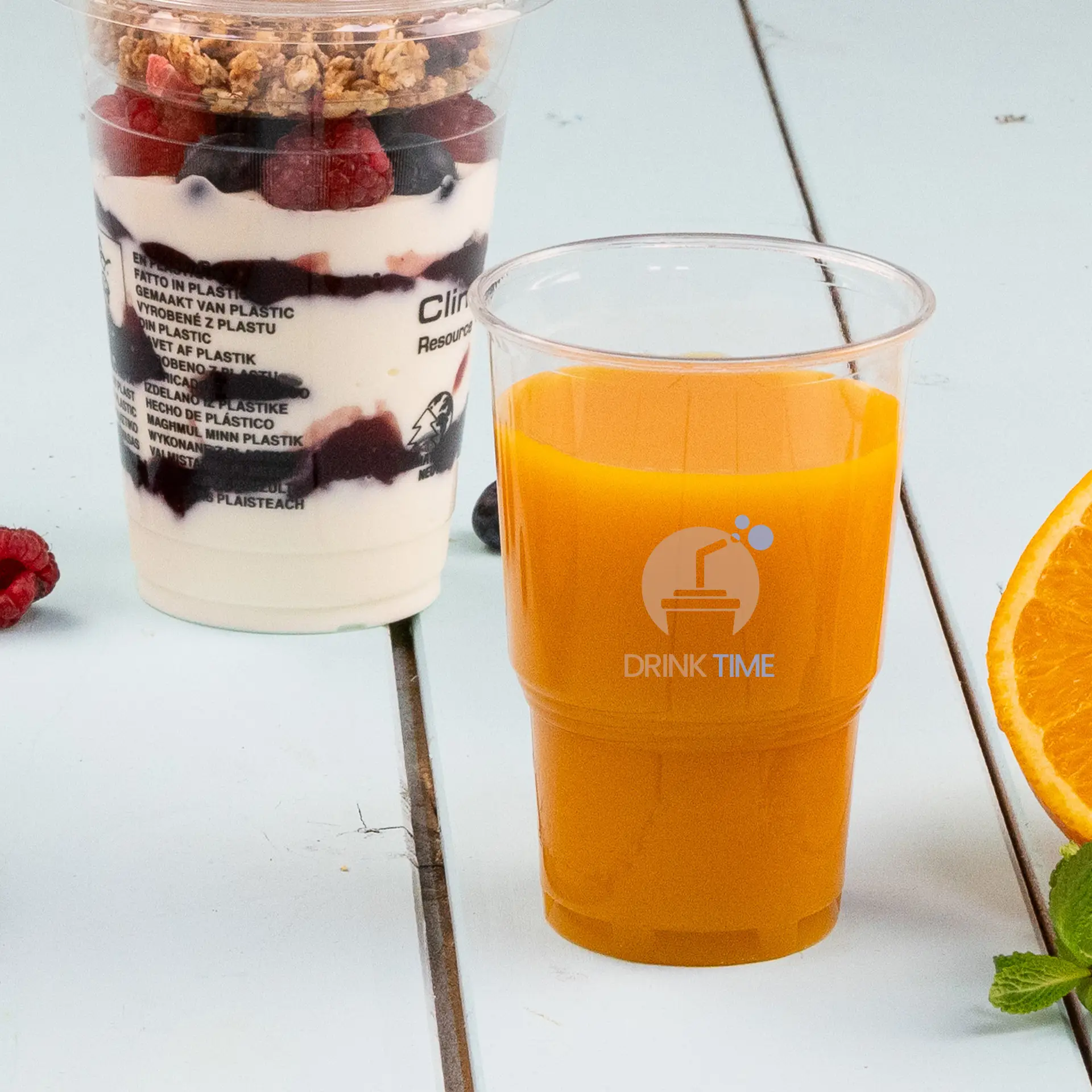 One clear 10 oz PLA cup with orange beverage and diamond graphic; sliced orange and mint nearby; parfait in background.