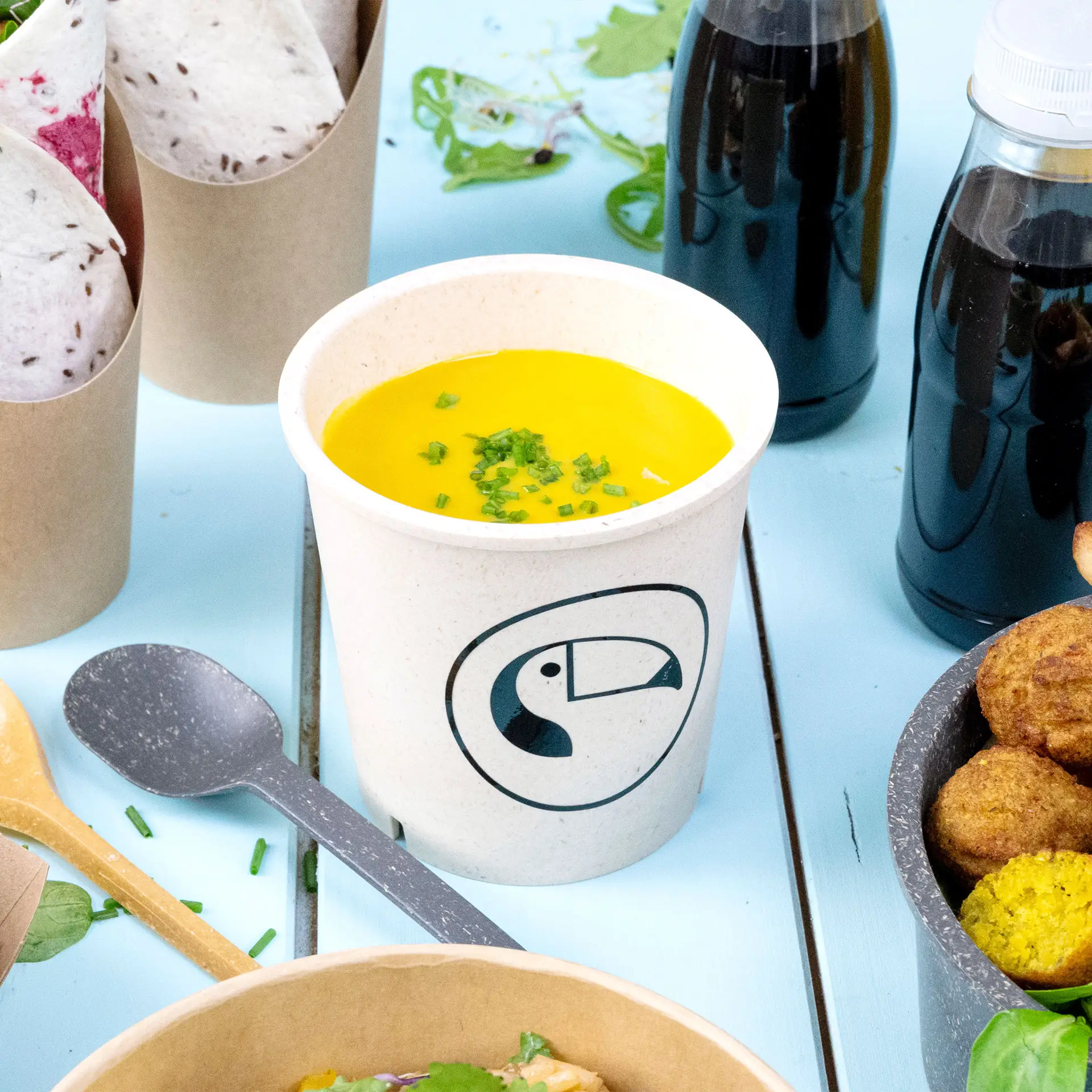 Cremefarbener, wiederverwendbarer Suppenbecher mit gelber Suppe und Schnittlauch, auf blauem Hintergrund mit anderen Lebensmitteln.