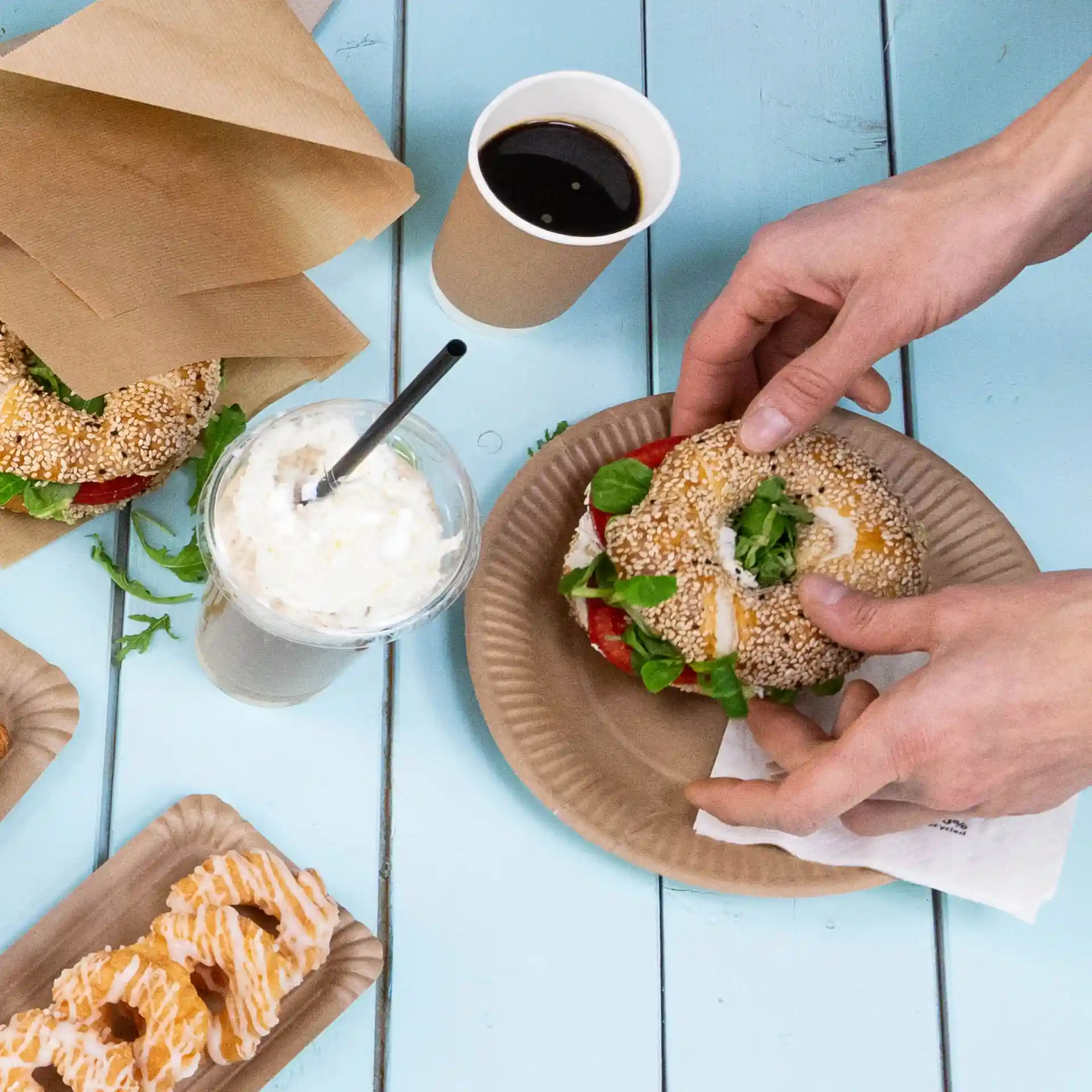 Image of a hand holding a sesame seed sandwich with lettuce and tomato on a brown paper plate, surrounded by a drink and pastries.