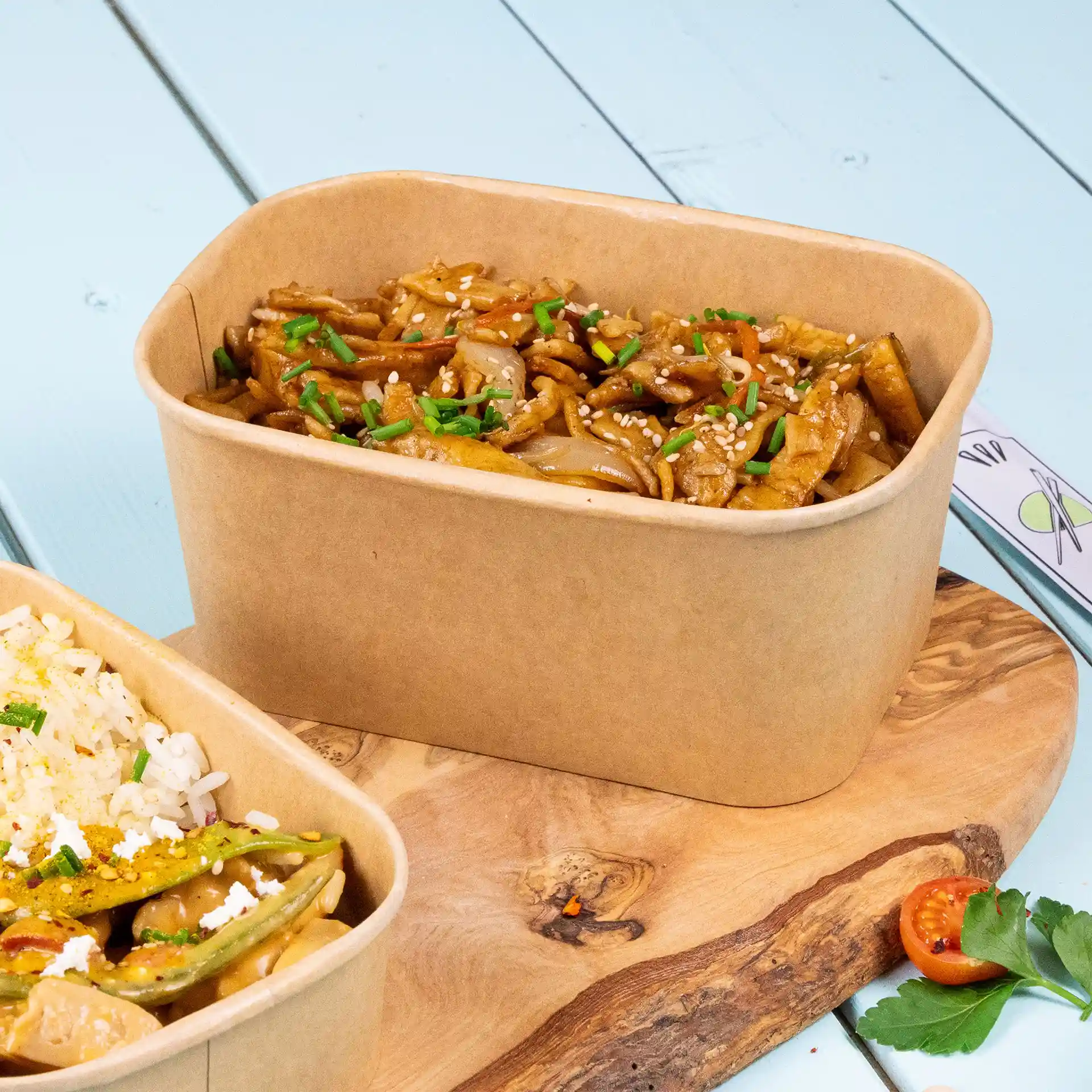 Rectangular kraft food container filled with noodles, topped with sesame seeds and chopped chives, on a wooden surface.