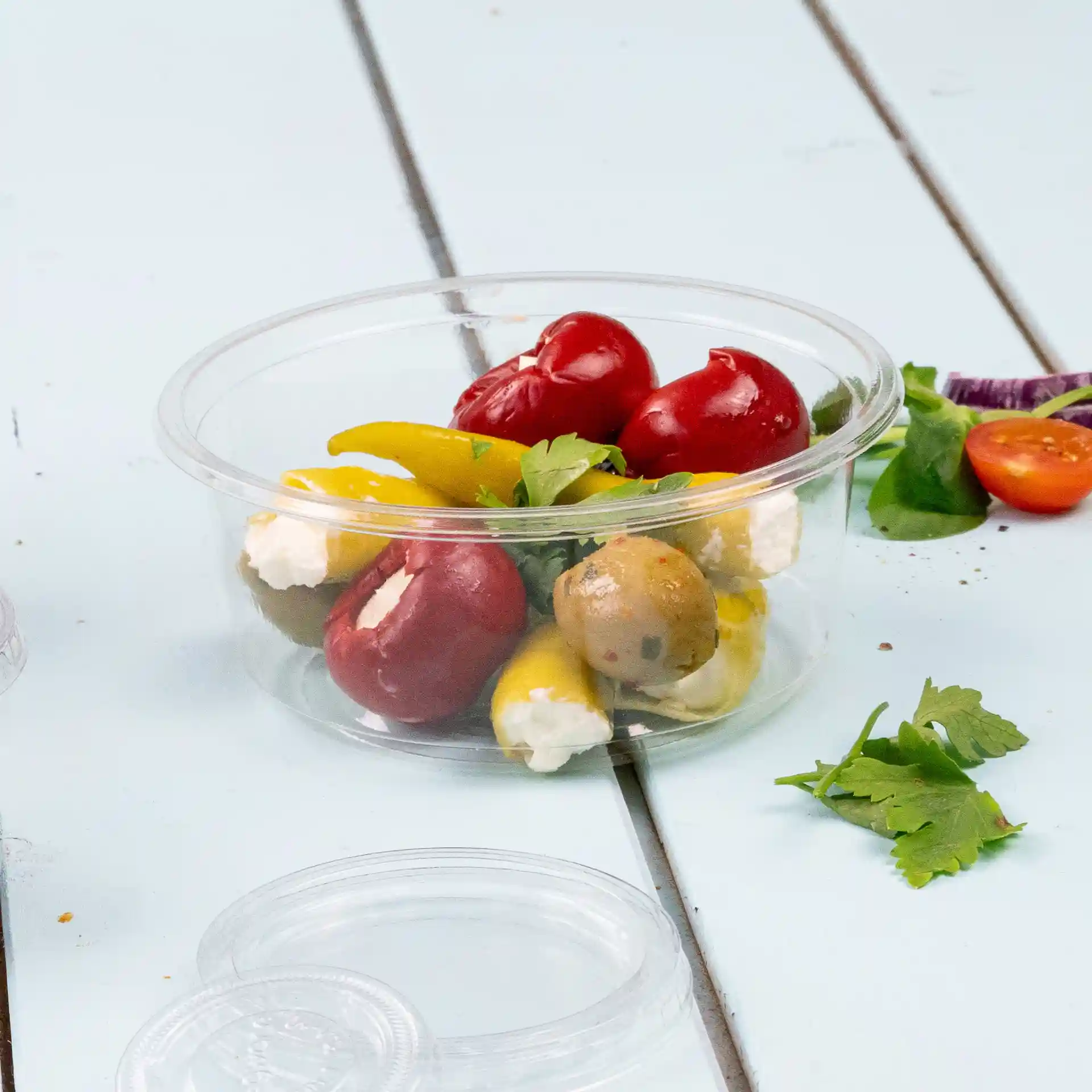 Transparenter rPET-Deli-Behälter mit verschiedenen Paprika, Oliven und Kräutern auf einer hellblauen Oberfläche.