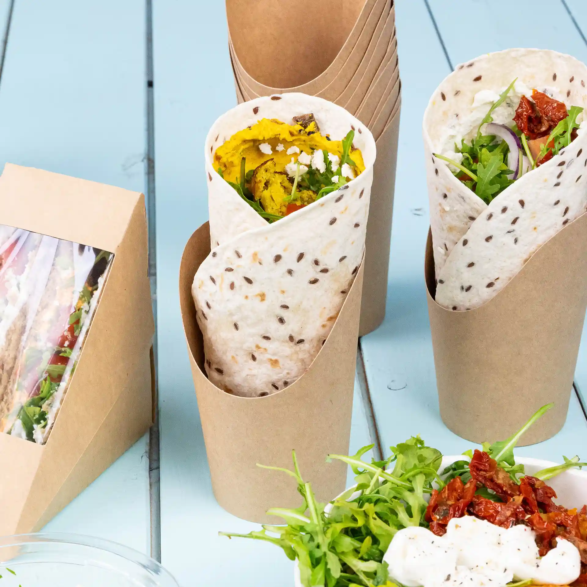 Bild zeigt zwei Wrap-Becher aus braunem Karton, einer gefüllt mit Essen und einem Beilagensalat mit grünen Blättern und getrockneten Tomaten.
