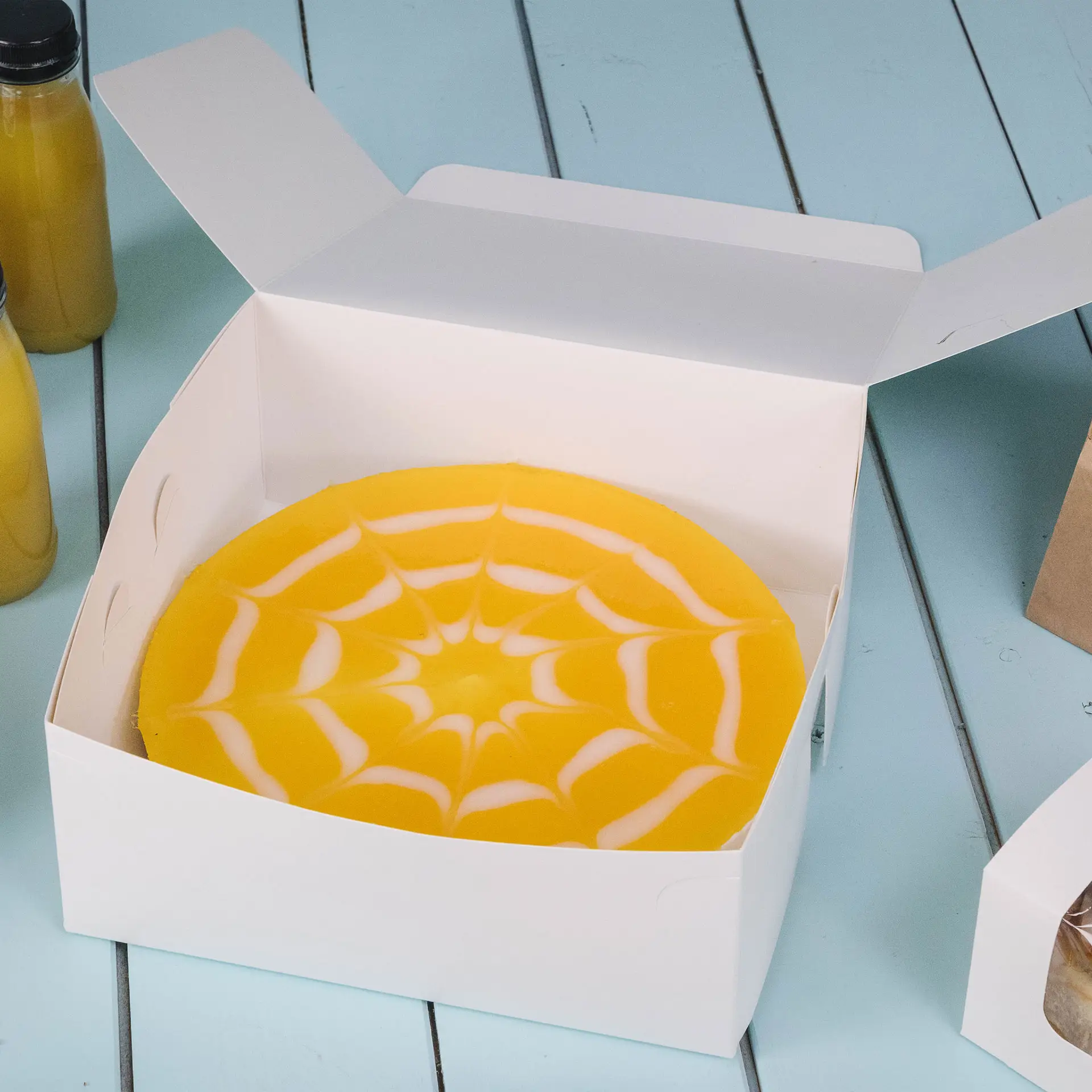 Weiße Kuchenbox mit einem gelben Kuchen mit Spinnennetzdesign innen, auf einer blauen Oberfläche platziert.