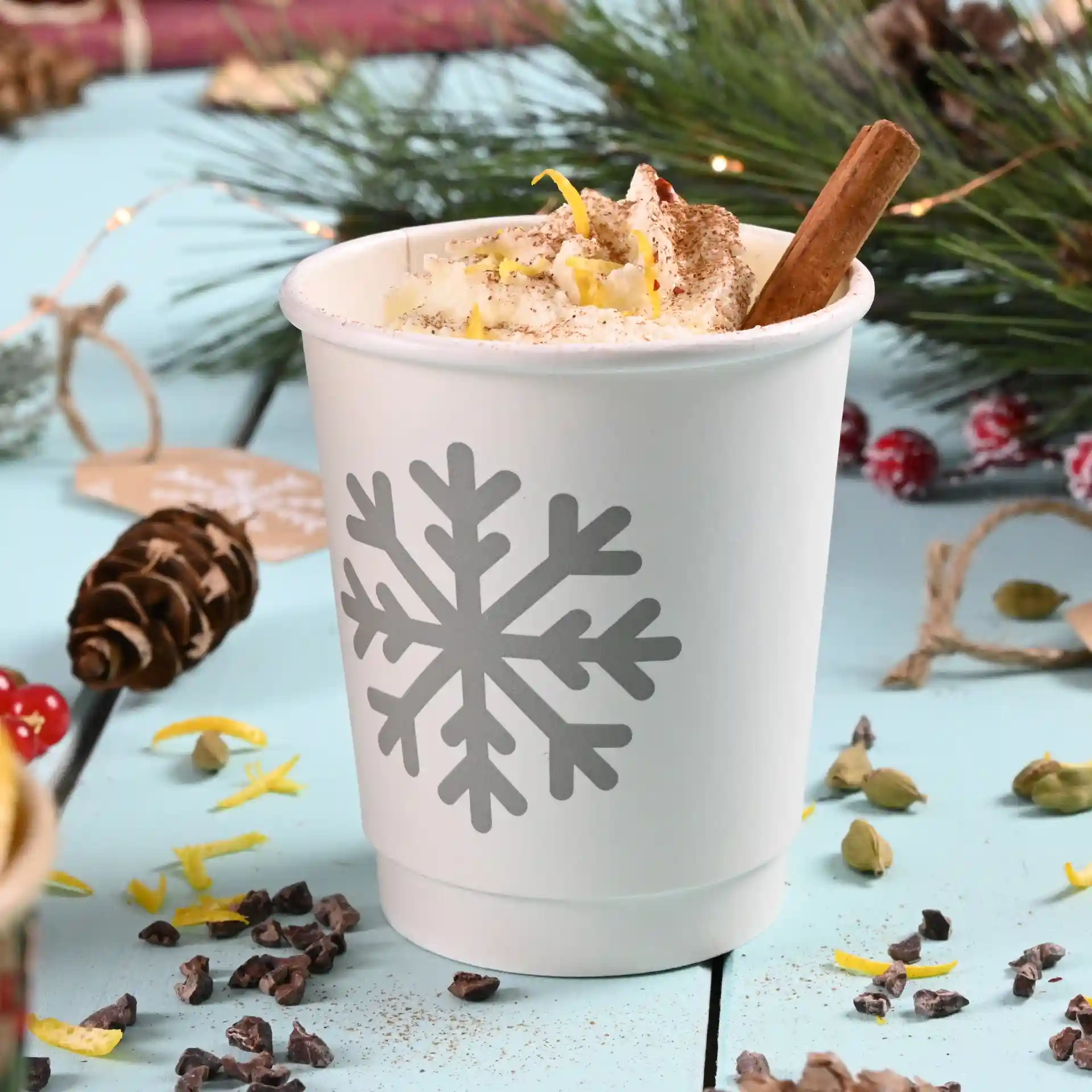 White double-walled paper cup with a grey snowflake design, filled with whipped cream and a cinnamon stick.