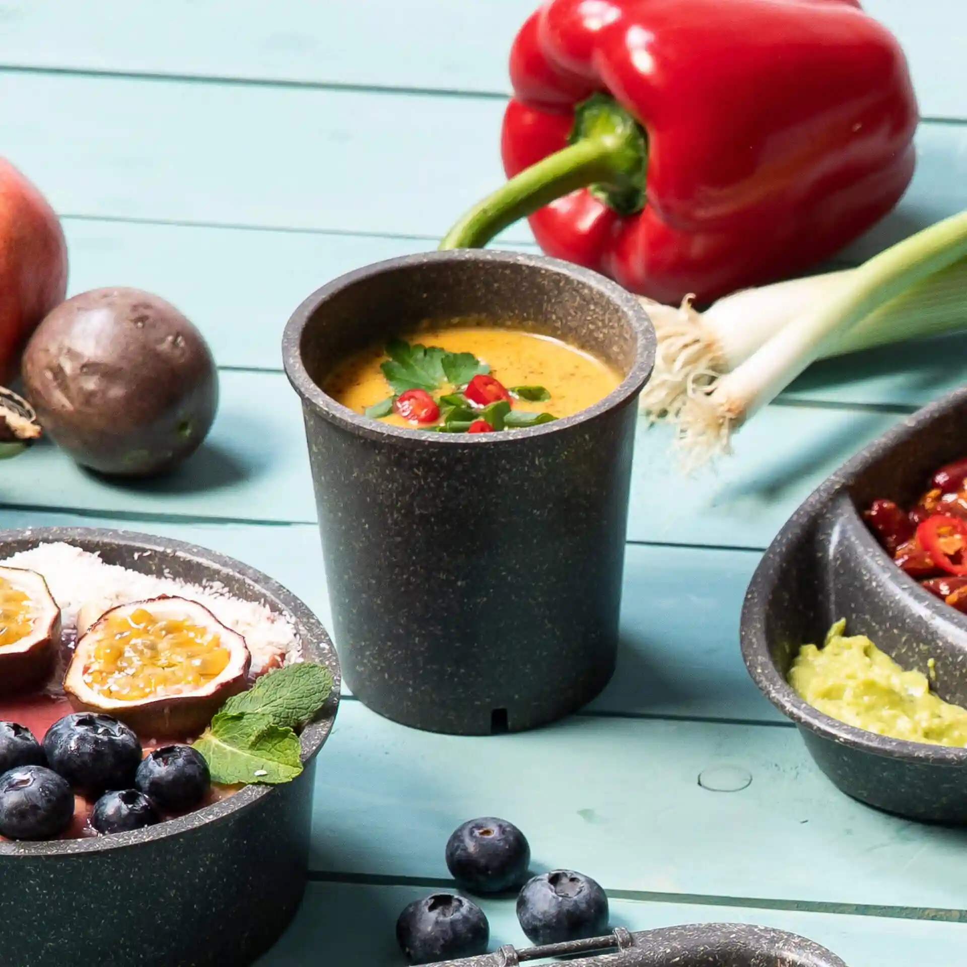 Grau wiederverwendbarer Becher mit Suppe, garniert mit Kräutern und Chili, umgeben von Obst und Gemüse auf einer blauen Fläche.