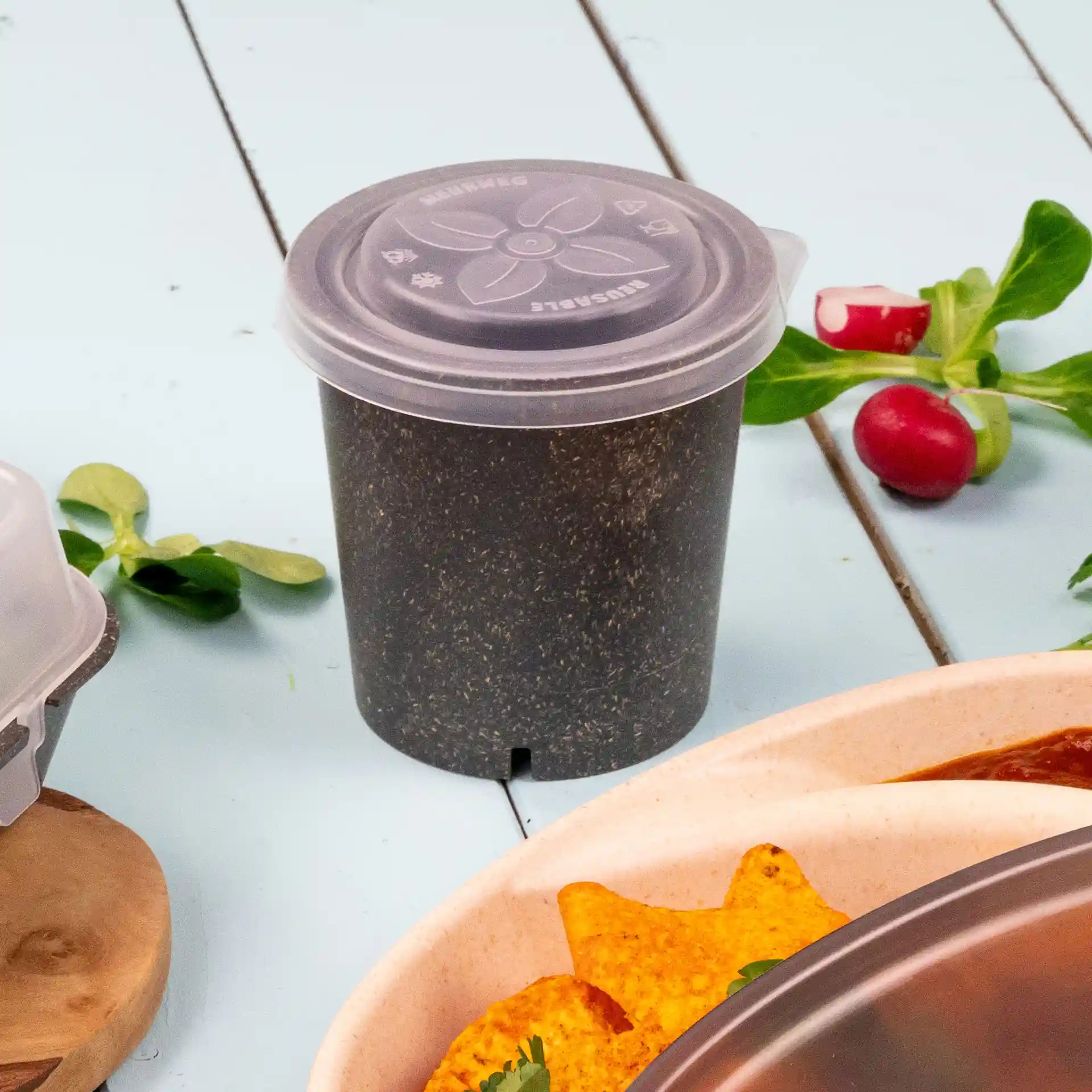 Transparent round cup with a lid, placed on a light blue surface, surrounded by green leaves and tortilla chips.