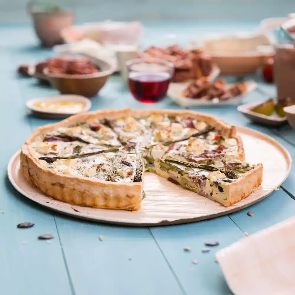 Savory quiche on a wooden plate, with a slice removed, surrounded by various dishes and a glass of red drink.