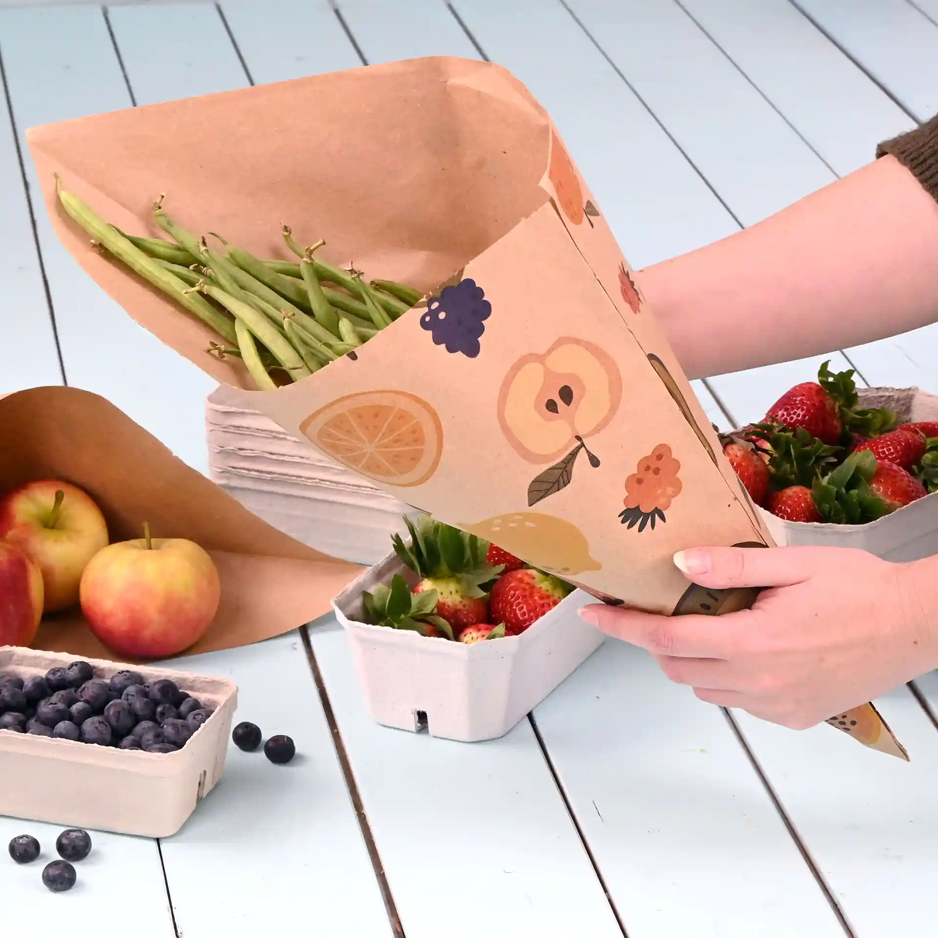 Papierbecher gefüllt mit grünen Bohnen, dekoriert mit Obstillustrationen, über Behältern mit Erdbeeren und Äpfeln gehalten.