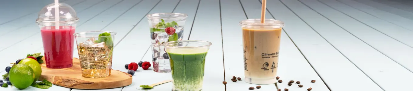 Verschiedene Getränke auf einer Holzoberfläche, darunter Eiskaffee, Smoothie, Joghurtparfait und Saft, mit einer Orangenscheibe daneben.