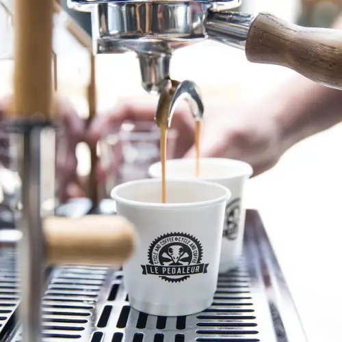 Coffee being brewed into two white cups with a logo labeled "Le Pedaleur."