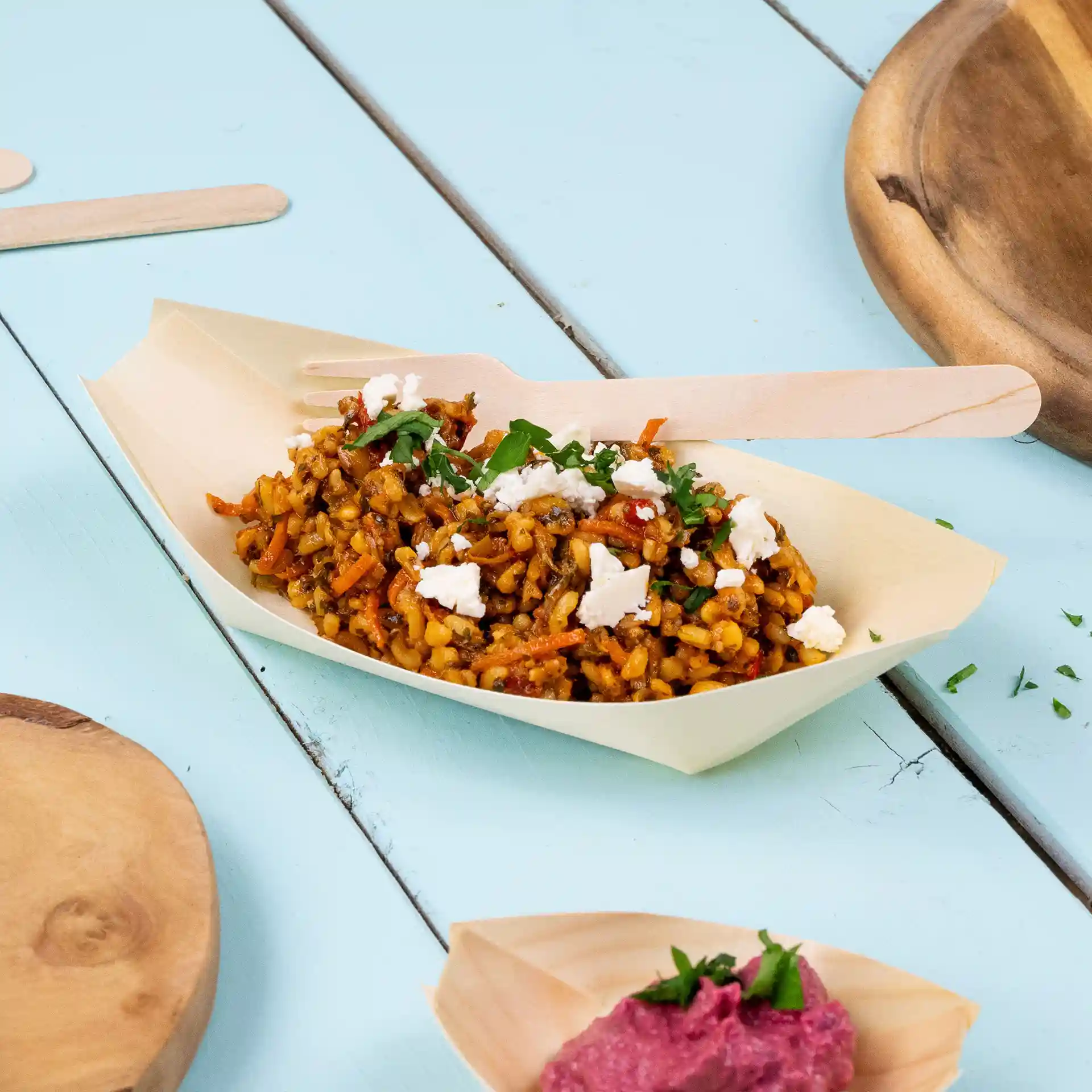 Holzboot-förmige Schale mit Reis, Gemüse und Feta-Käse, begleitet von einer Holzgabel auf einer blauen Oberfläche.