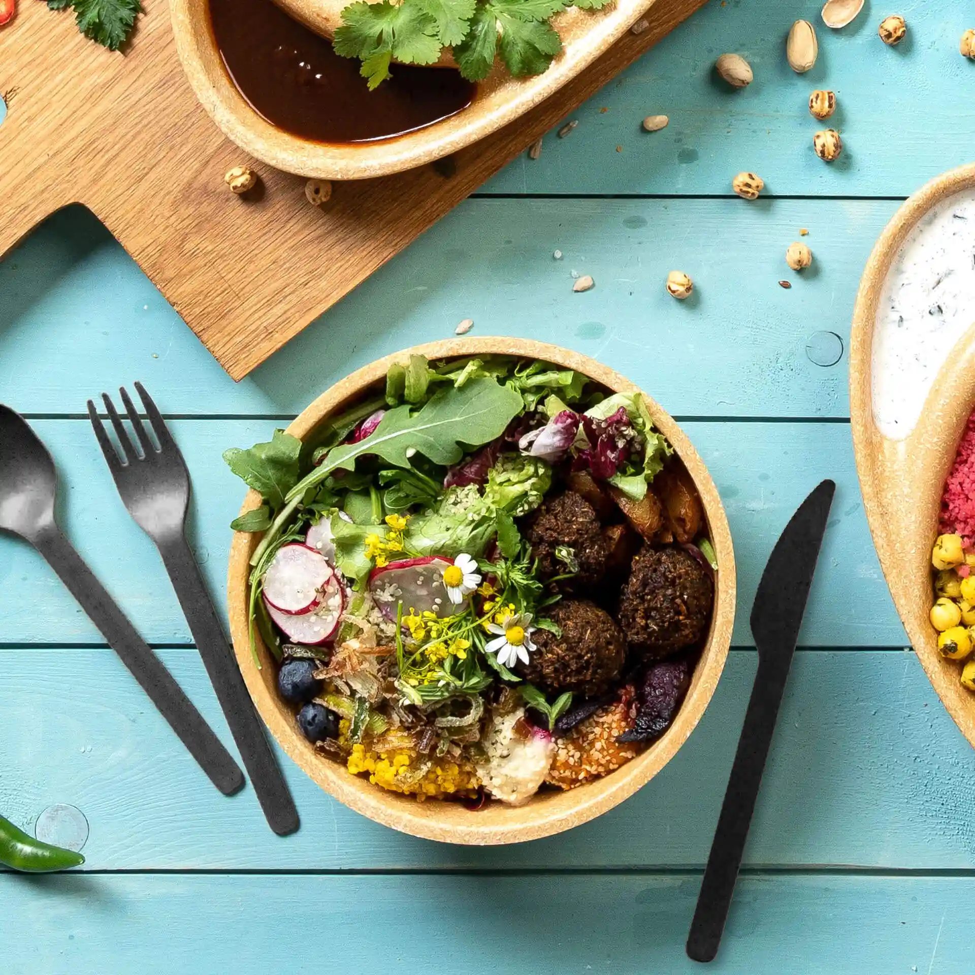Bowl filled with salad, falafel, and edible flowers, accompanied by black reusable cutlery on a blue wooden surface.