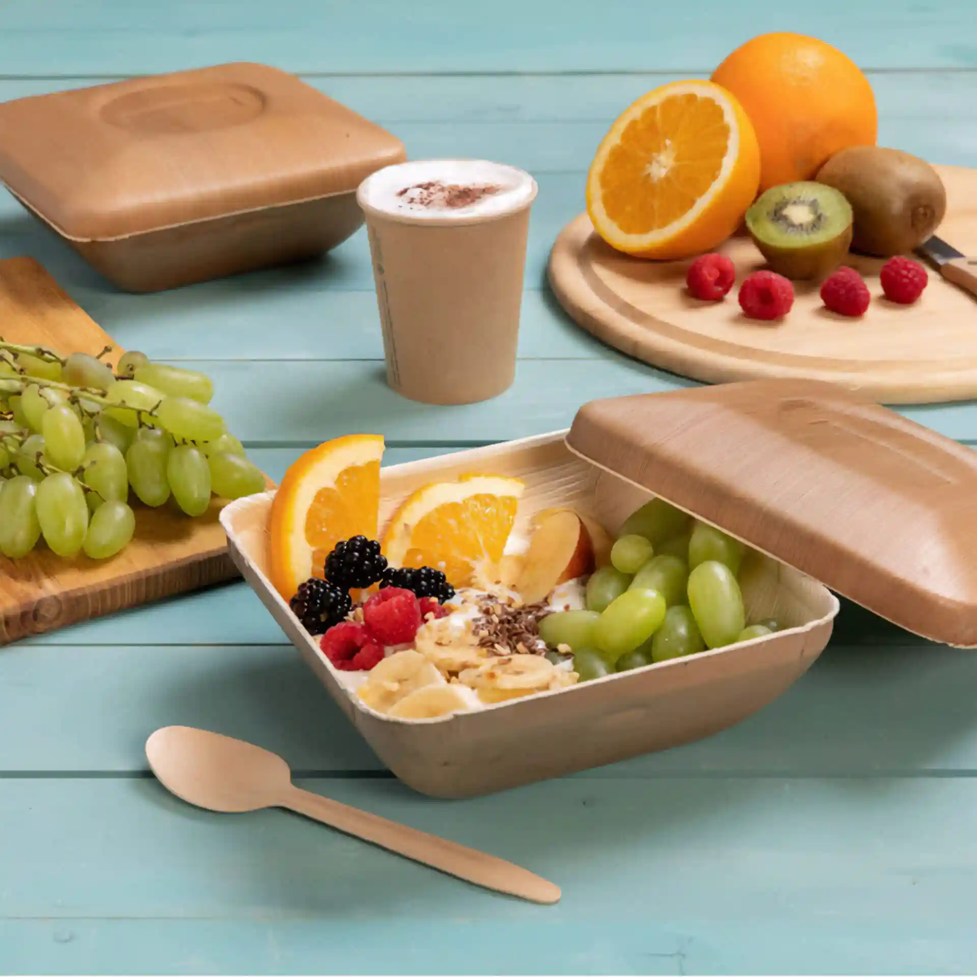 Quadratische Palmblatt-Schüssel mit Deckel, gefüllt mit Obst, Löffel und einem Becher auf einer blauen Holzoberfläche.