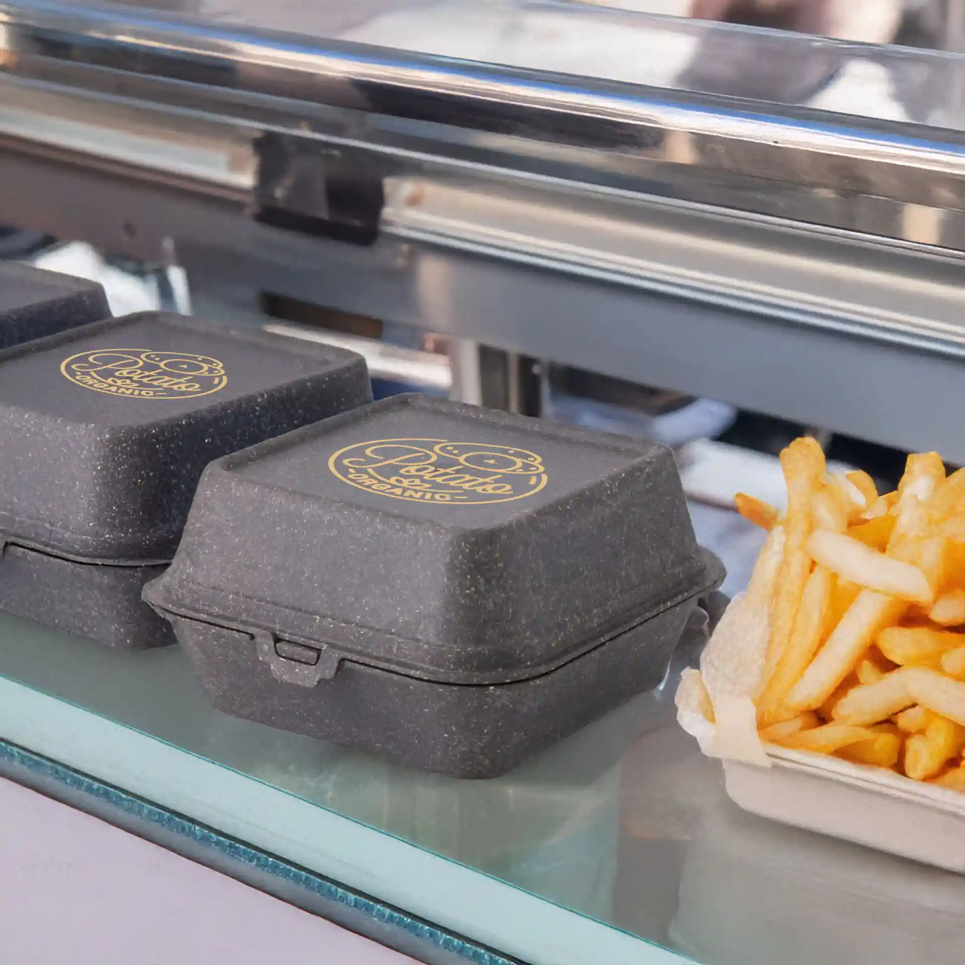 Drei graue bedruckte wiederverwendbare Burgerboxen mit einem Logo oben, neben einer Portion Pommes frites platziert.
