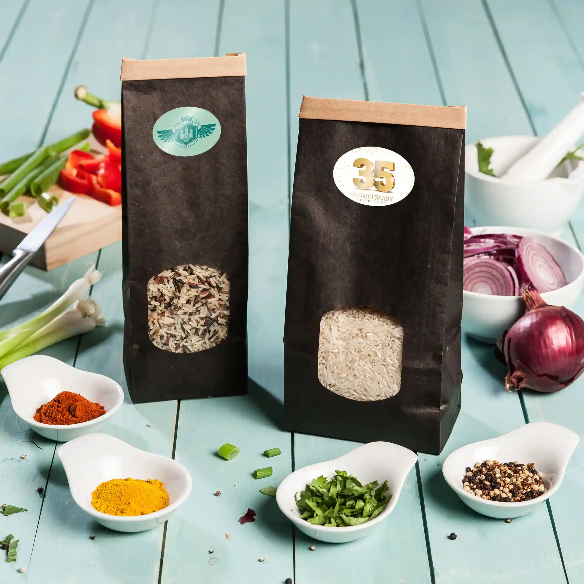 Two black paper bags containing rice, with visible labels, surrounded by various spices and chopped vegetables on a blue wooden surface.