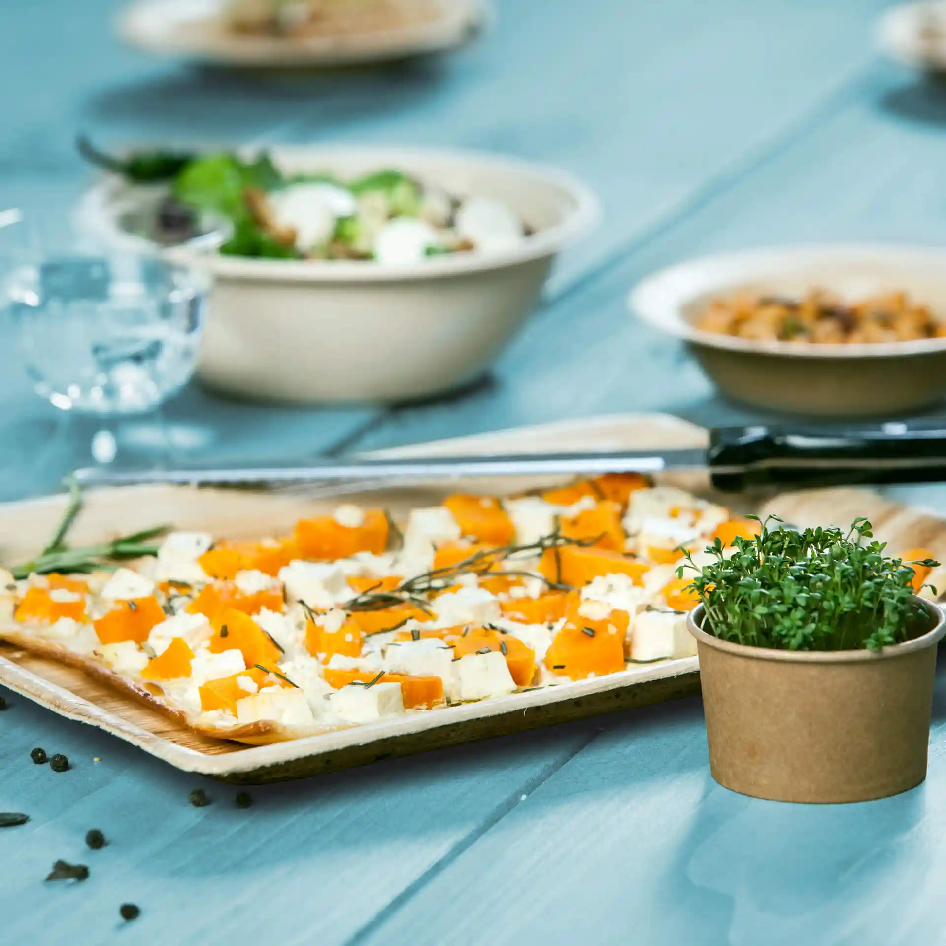 Rechteckiges Palmblatt-Tablett mit orange-weißen Lebensmittelwürfeln, garniert mit Kräutern, auf einem blauen Holztisch.