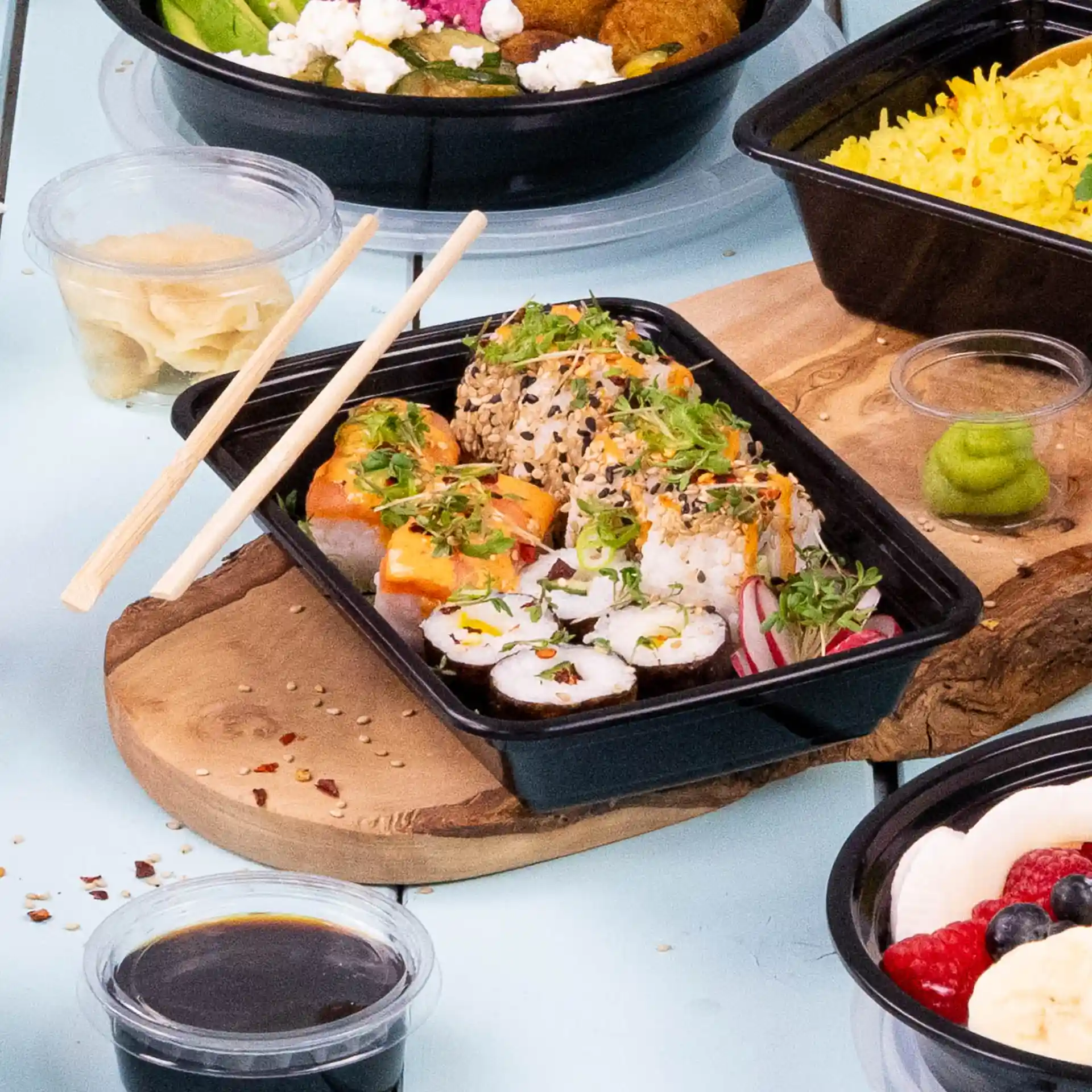 Sushi rolls with toppings in a black rectangular reusable container, chopsticks beside, and condiments including wasabi and soy sauce.