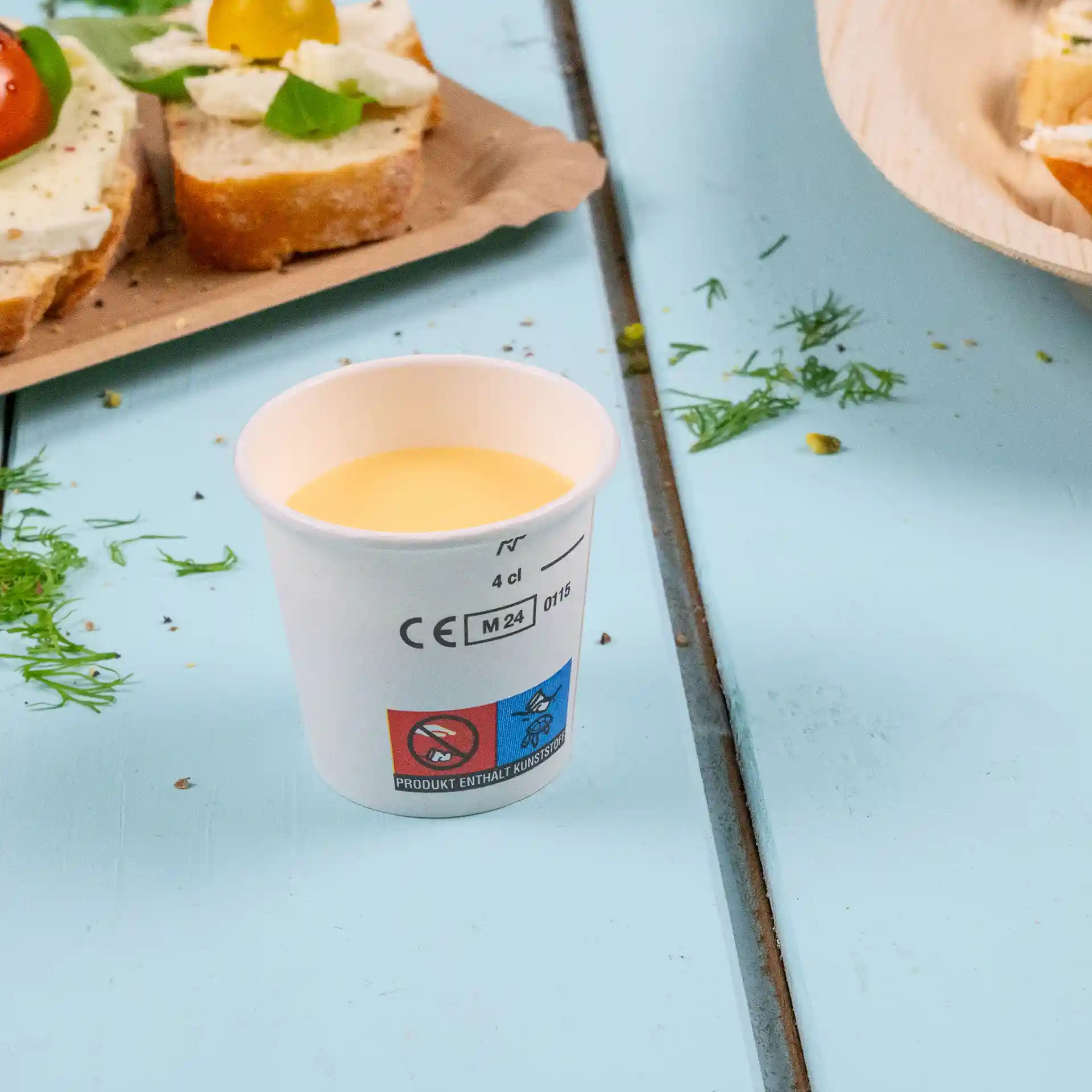 Weißer Becher mit gelblichem Getränk, auf blauer Fläche mit Lebensmitteln im Hintergrund.