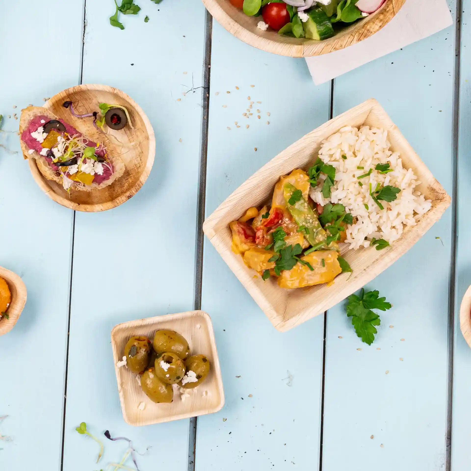 Image of various food items served in palm leaf bowls on a blue wooden surface, including rice, olives, and a salad.