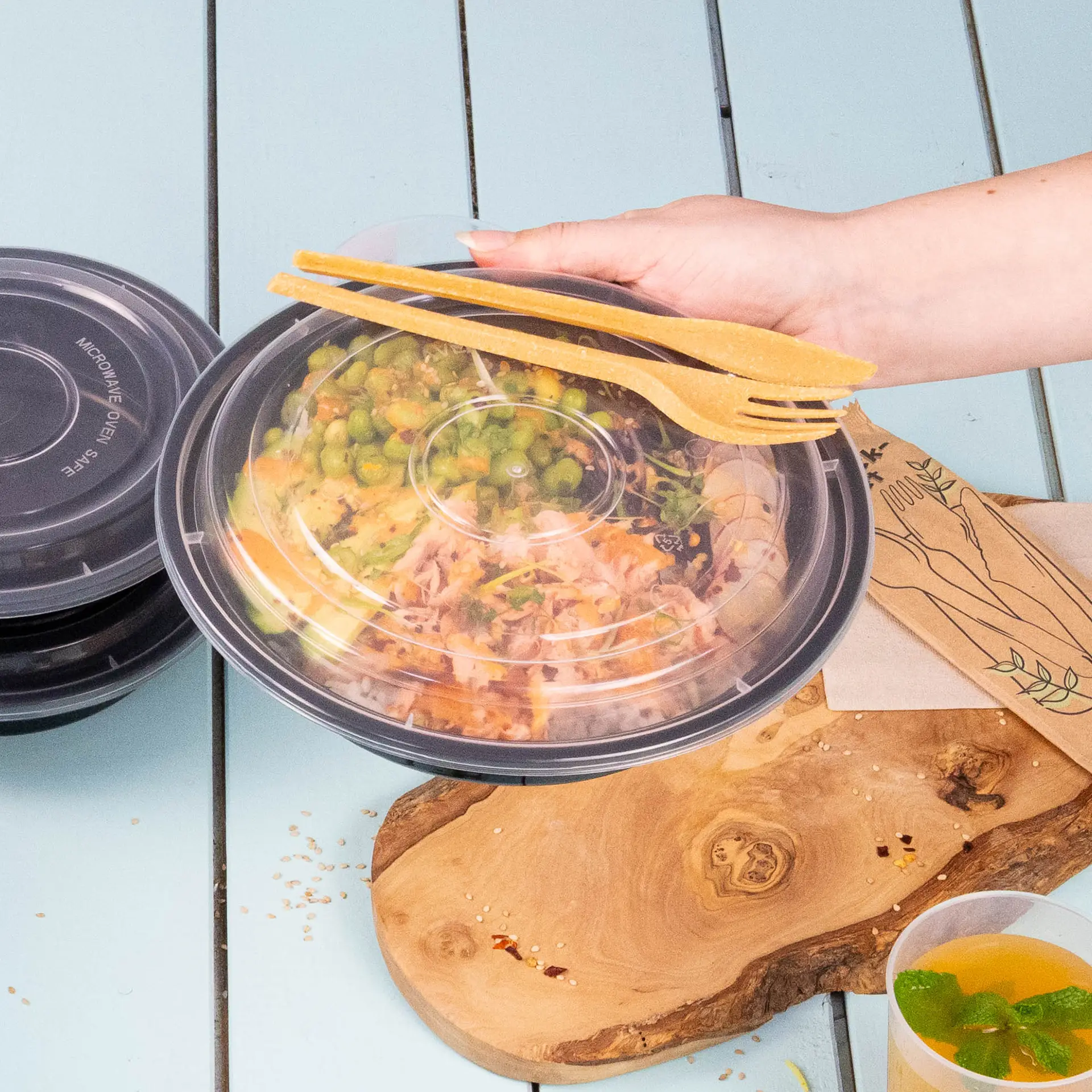 A hand holds a transparent, reusable lid over a bowl of colorful food; multiple black containers are nearby, complementing the scene.