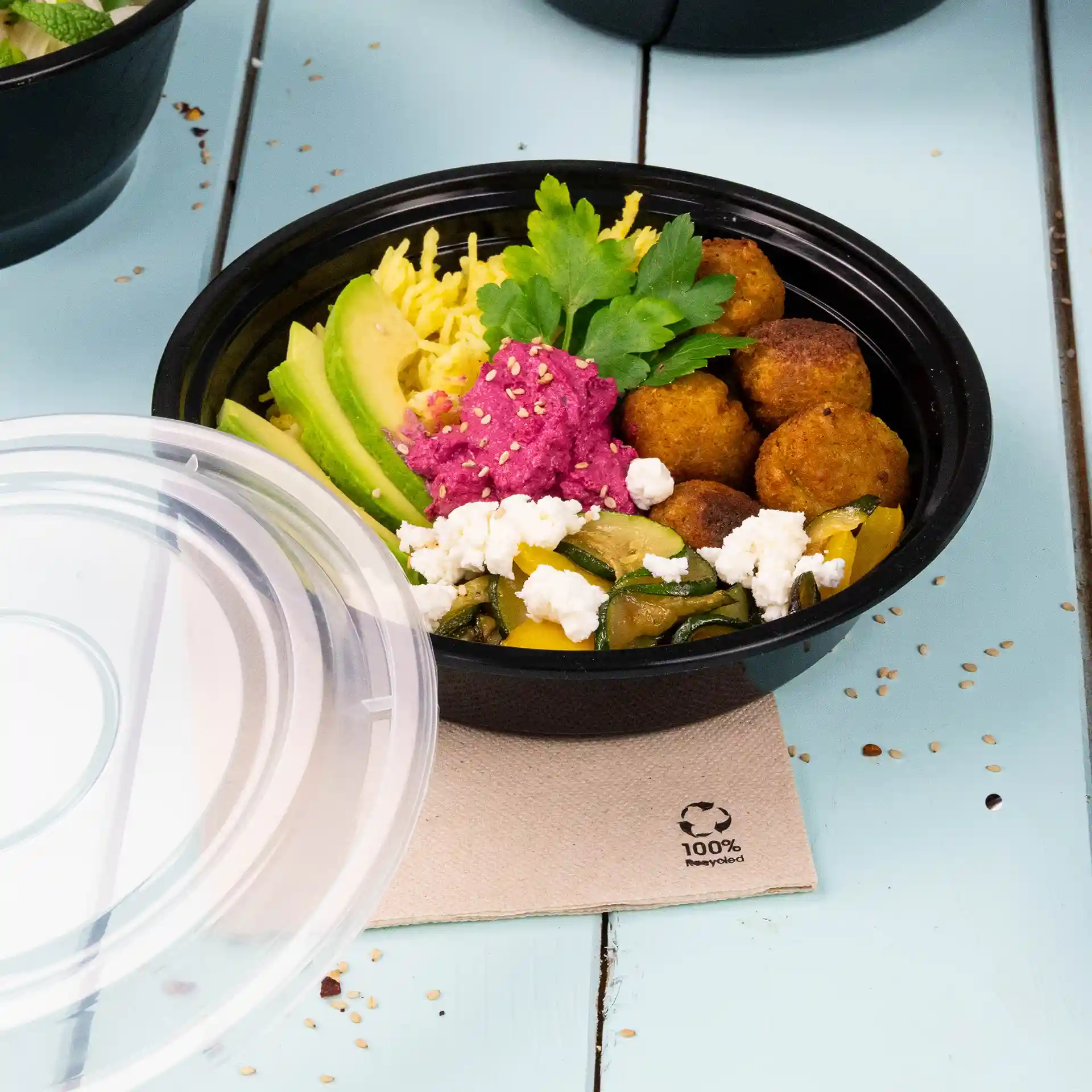 Schwarzer runder wiederverwendbarer Teller mit verschiedenen Lebensmitteln, darunter Avocado, Falafel und einem rosa Aufstrich, auf einer hellblauen Oberfläche.