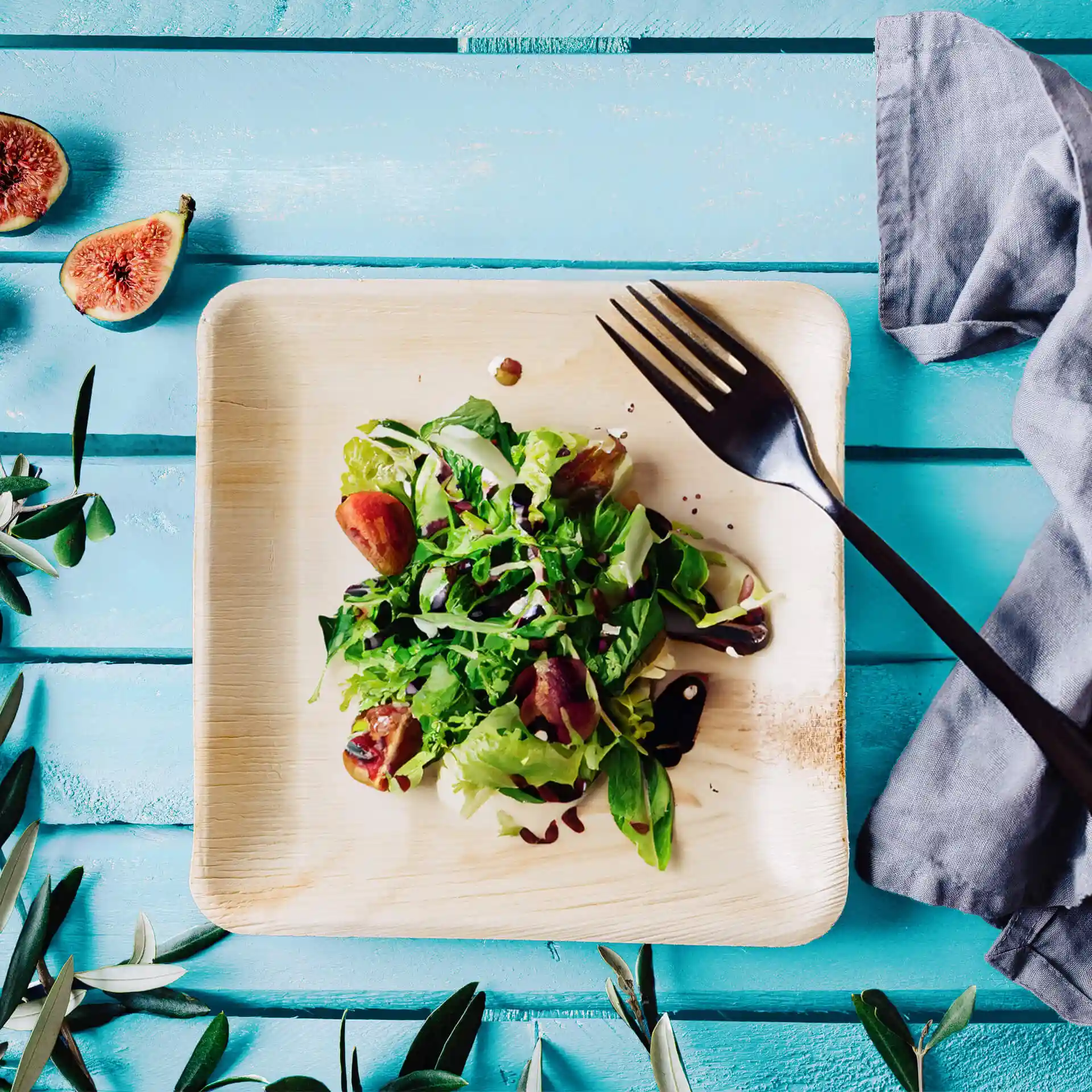 Quadratische Palmblattplatte mit einem Salat, garniert mit Grün und Feigen, dazu eine schwarze Gabel auf einer blauen Holzoberfläche.