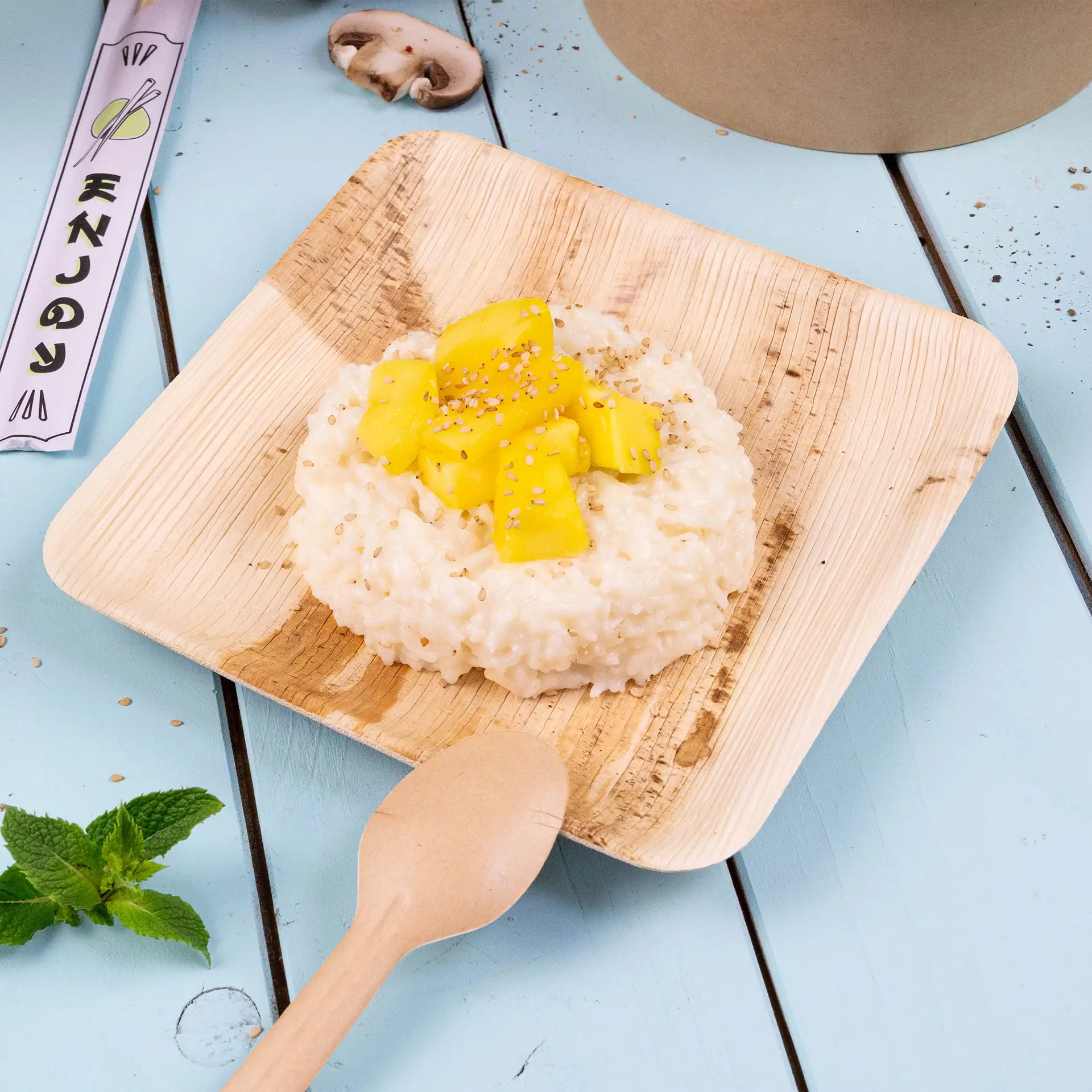 Square palm leaf plate with risotto topped with mango and sesame seeds, accompanied by a wooden spoon and mint leaves.