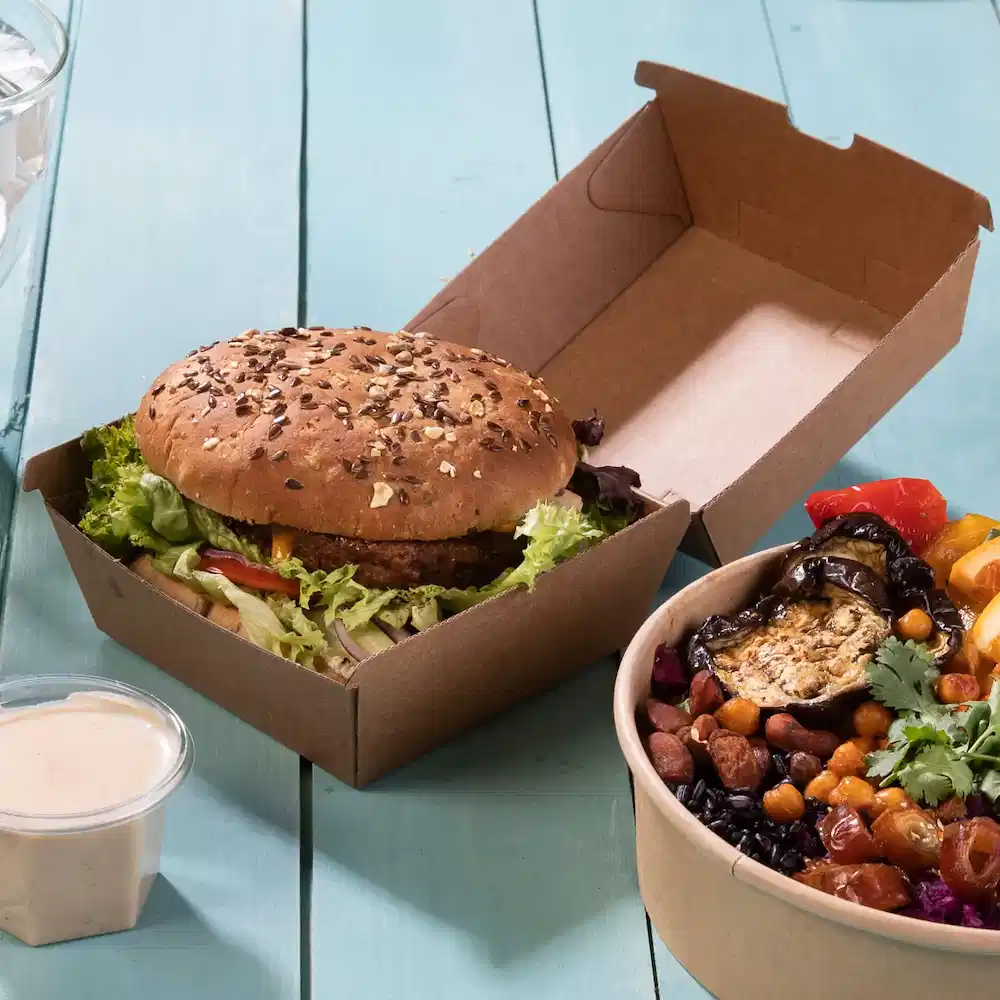 Ein Burger mit Sesambrötchen und Salat in einer braunen Takeaway-Box neben einer Schüssel mit gemischtem Gemüse und Hülsenfrüchten.