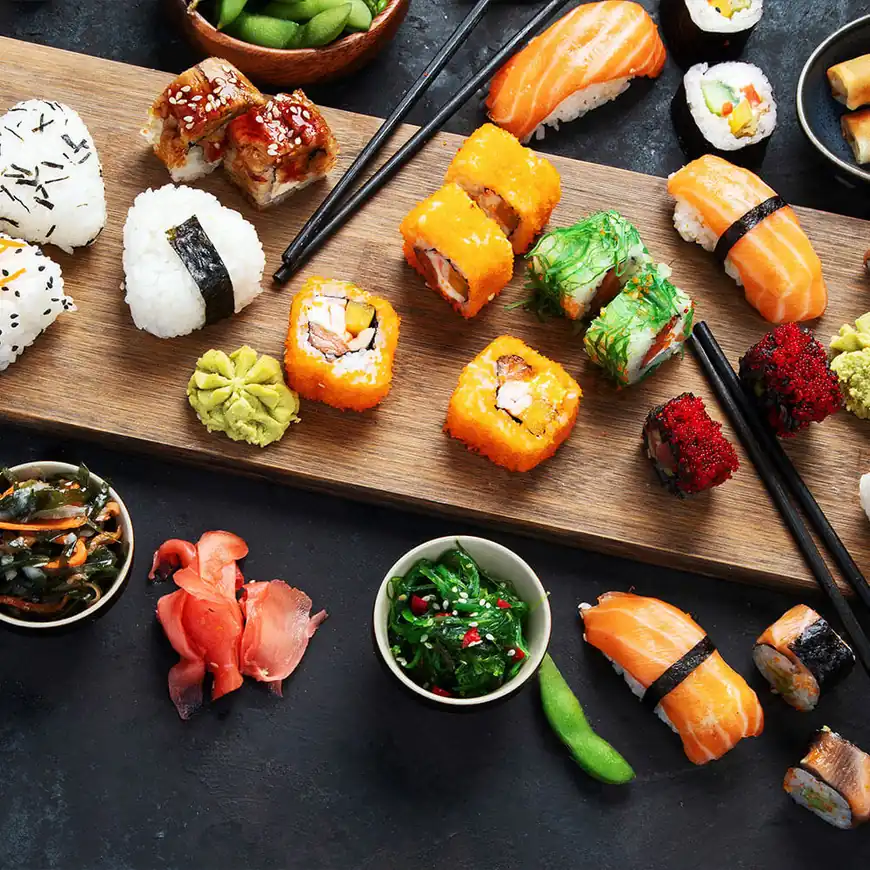 Ein Holztablett mit verschiedenen Sushi-Arten, darunter Rollen, Sashimi und Nigiri. Begleitet von Wasabi, eingelegtem Ingwer und kleinen Schalen mit Algensalat.