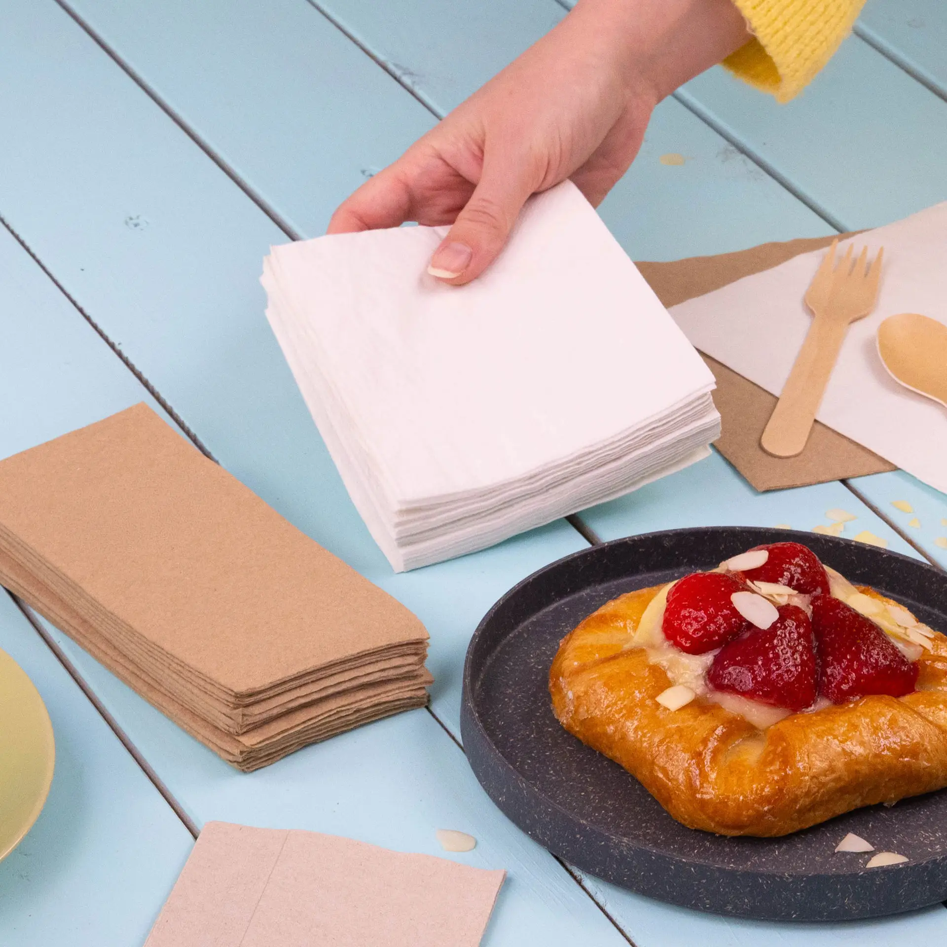 White 2-ply cocktail napkins in a stack, with a hand reaching for them, alongside a pastry on a plate.