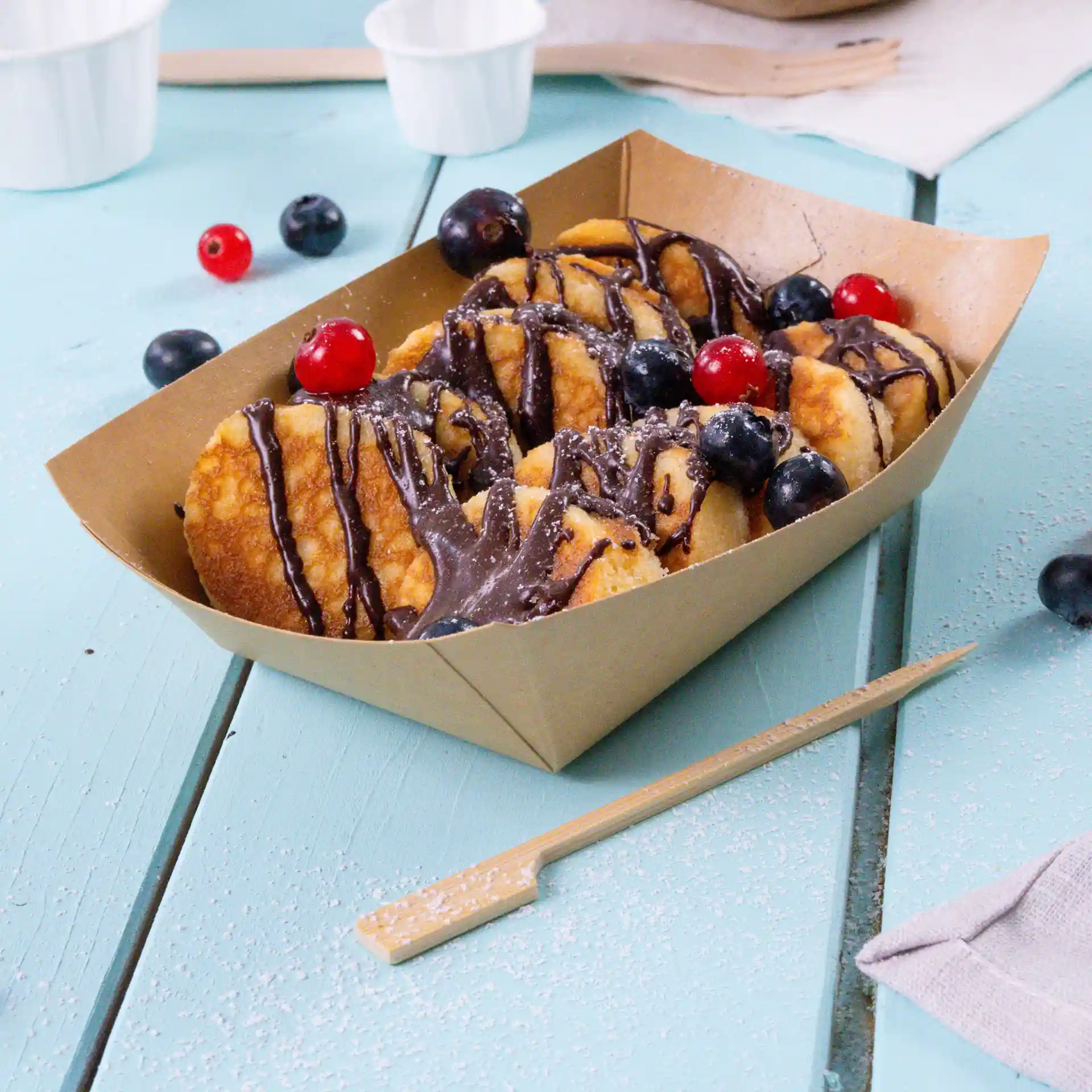 Brown cardboard snack tray filled with dessert, topped with chocolate drizzle, blueberries, and red currants.