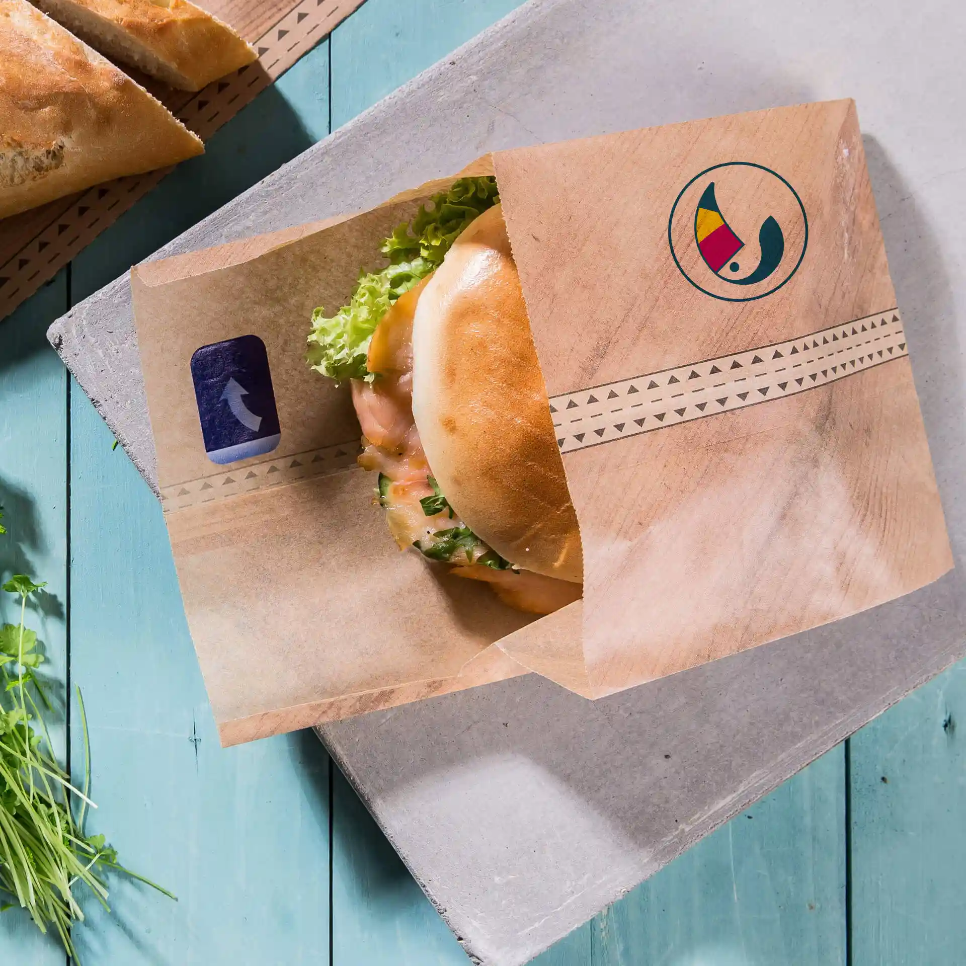 Braune, individuell bedruckte Burgerhülle mit Klebeverschluss, die einen Burger mit Salat auf einer blauen Holzoberfläche hält.