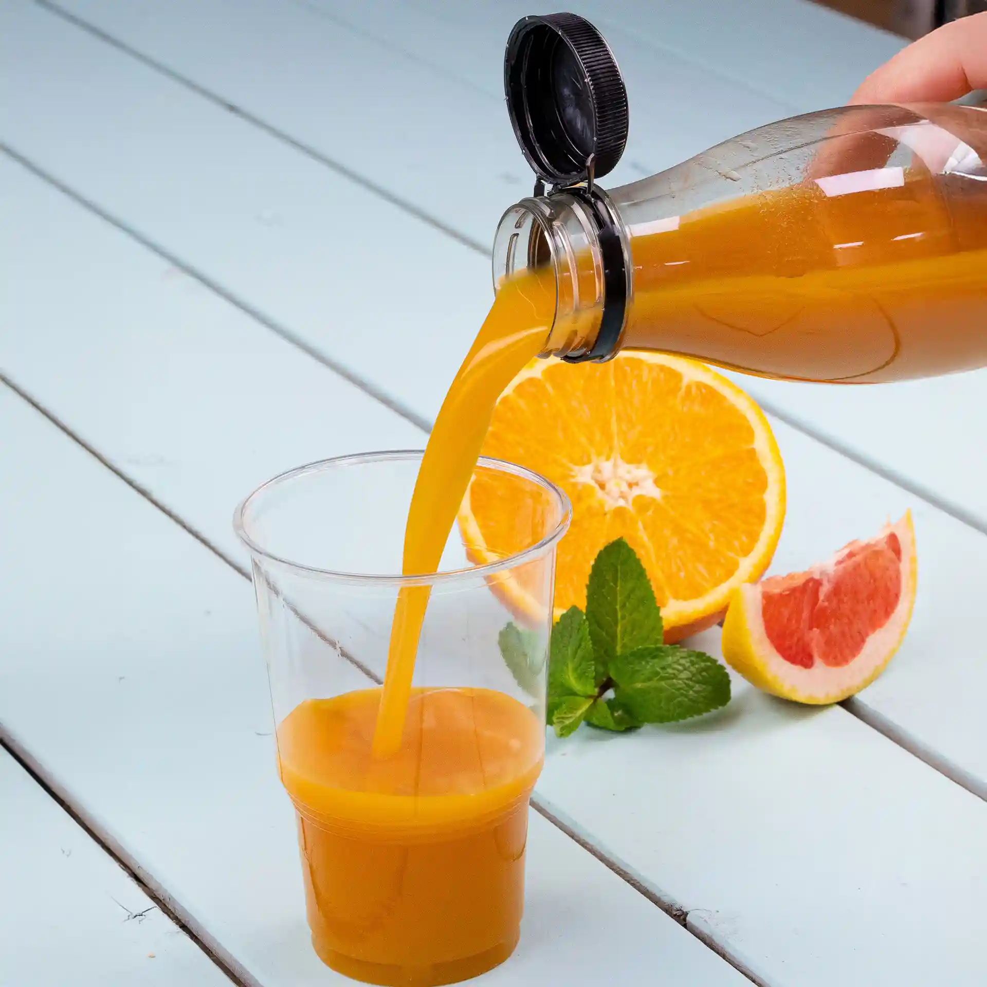 Transparente rPET-Flasche, die Orangensaft in einen klaren Becher gießt, mit Orangen- und Grapefruitscheiben daneben.