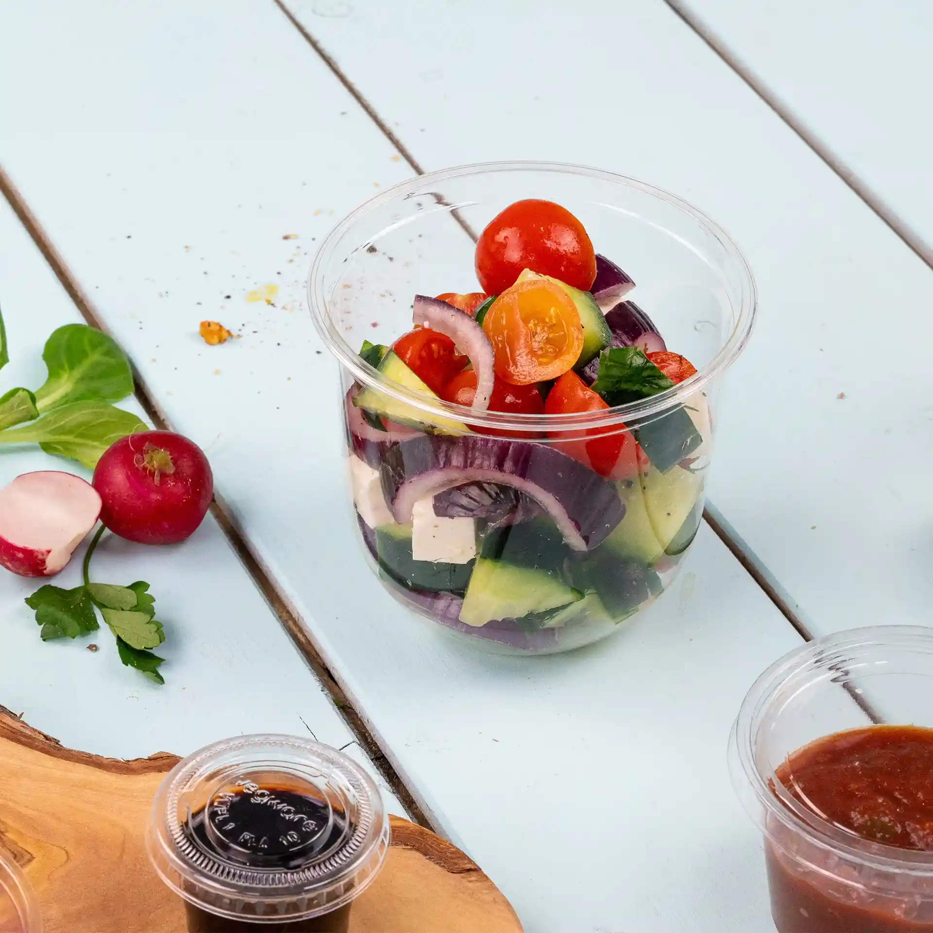 Transparenter Becher gefüllt mit gemischten Salatzutaten, darunter Tomaten, Gurken und Zwiebeln, auf einer hellblauen Oberfläche.