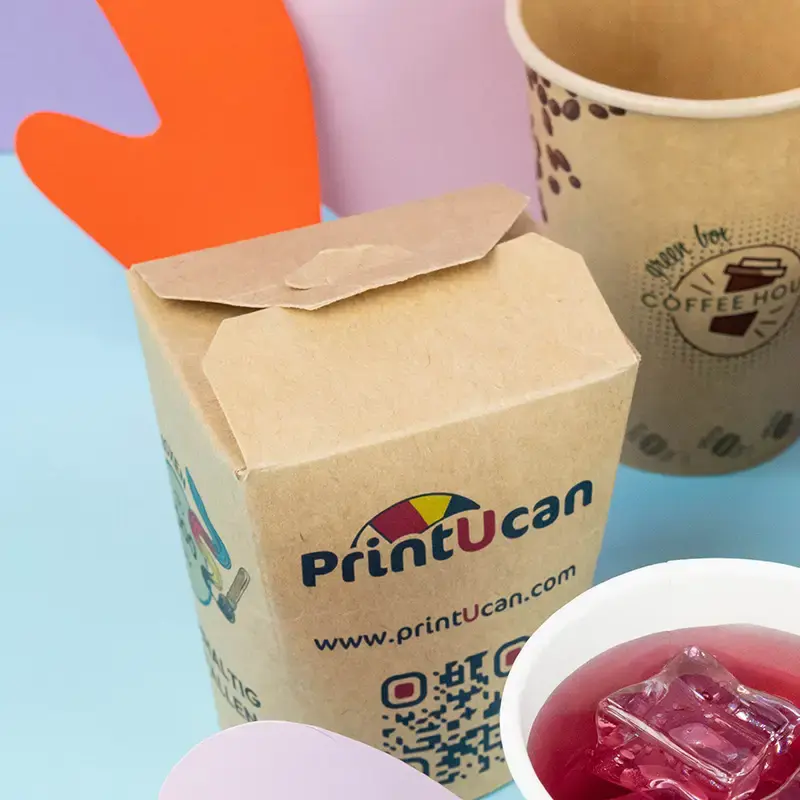 Brown cardboard containers for ramen noodles with a logo, placed on a blue surface with various toppings and chopsticks.