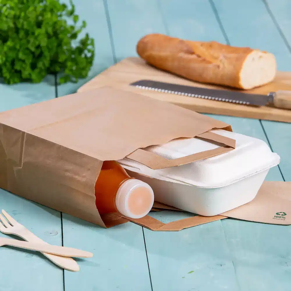 Braune Papiertüte mit einer Getränkeflasche und einem Takeaway-Essensbehälter auf einer blauen Holzoberfläche.