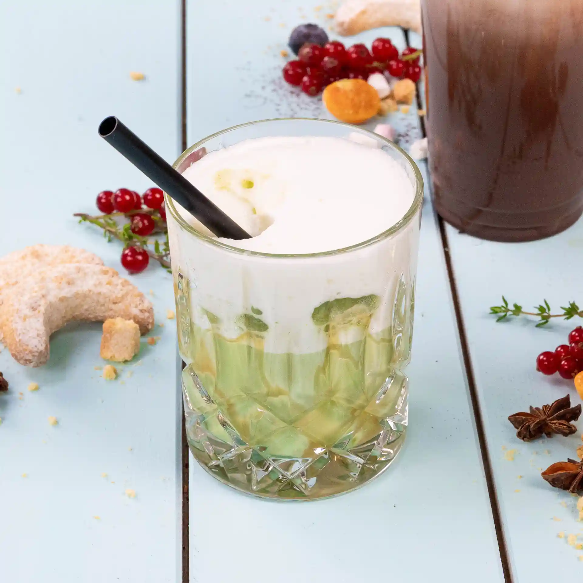 Glass with layered drink topped with foam and a black straw, surrounded by cookies and berries on a blue surface.