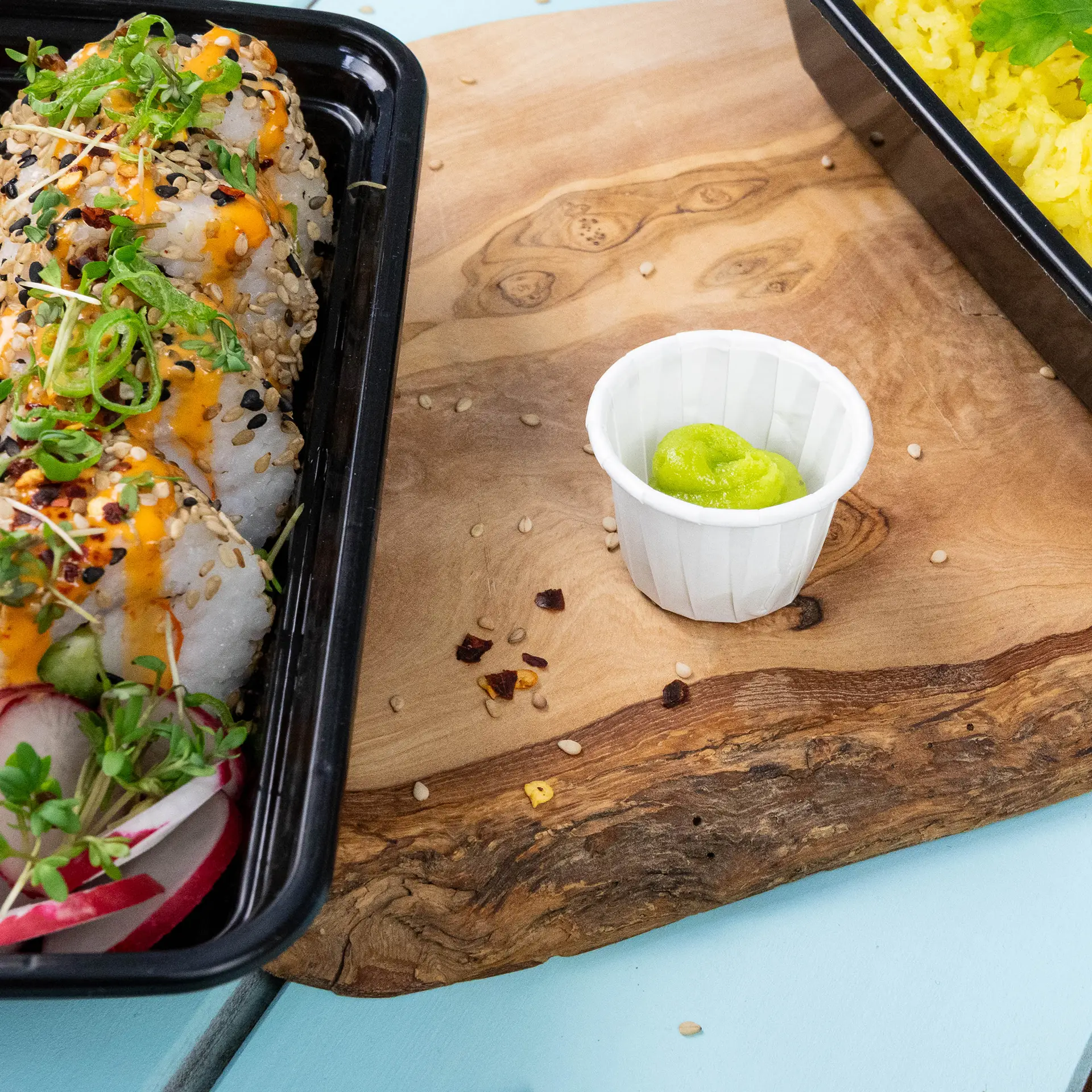 Black container with sushi rolls topped with sauce and sesame seeds, beside a small white paper cup of wasabi on a wooden surface.