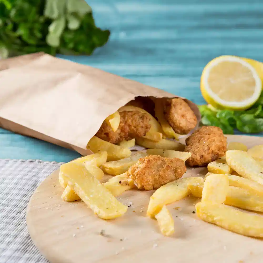Eine Papiertüte enthält frittierte Hühnchen-Nuggets und goldene Pommes, mit einer Zitronenscheibe und Gemüse im Hintergrund.