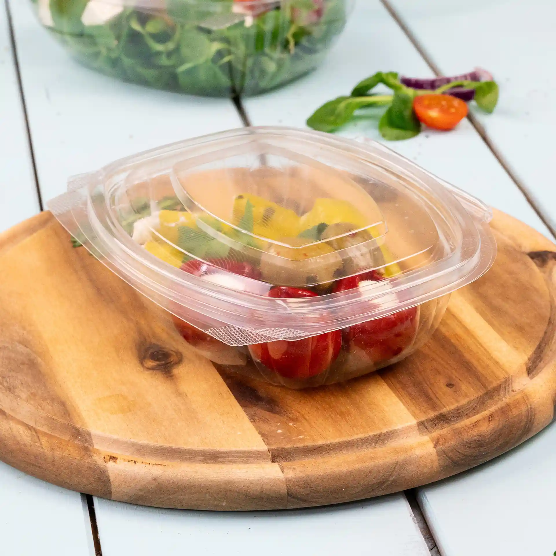 Transparent rectangular PLA deli bowl with a folding lid, containing mixed vegetables, placed on a wooden cutting board.