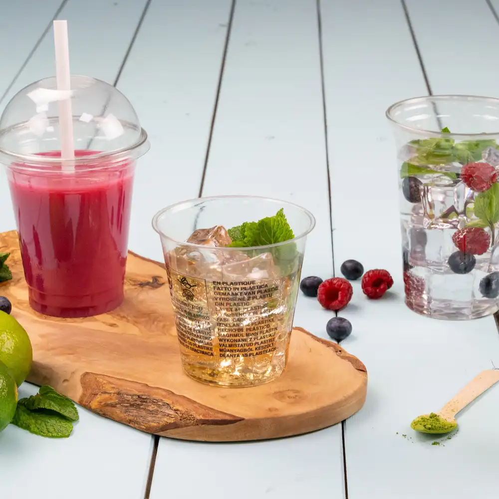 Clear plastic cups with drinks and mint placed on a wooden board, with berries and lime scattered around.