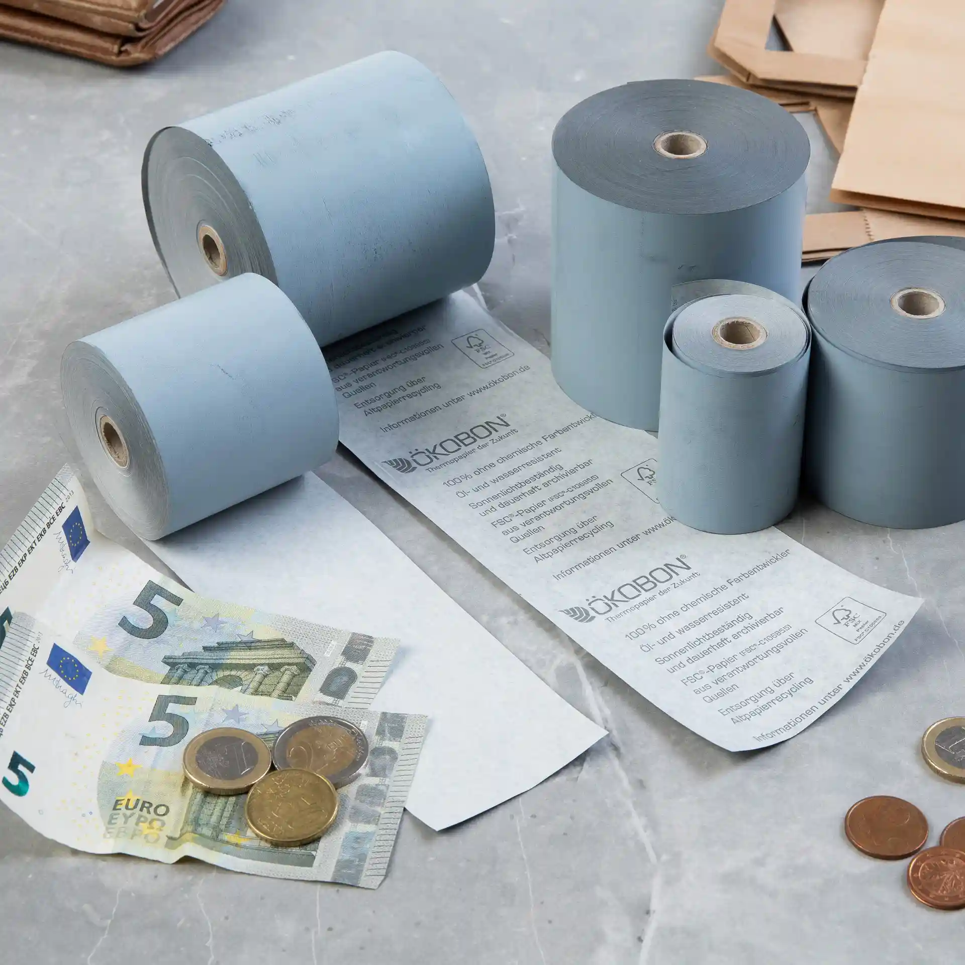 Image of several rolls of grey thermal paper, a 5 euro banknote, and coins on a surface.