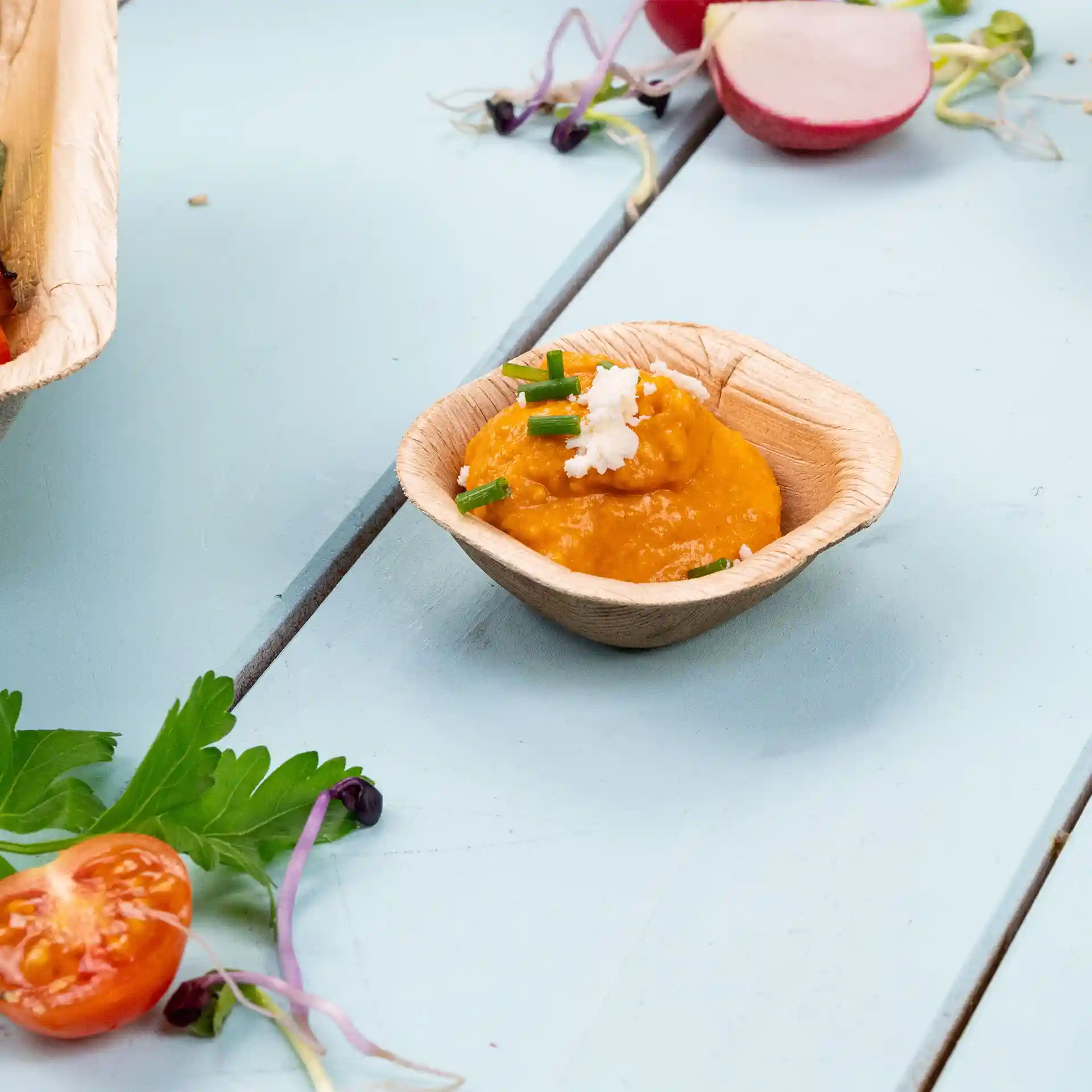 Deep square palm leaf bowl containing orange dip topped with chives and cheese, placed on a light blue surface.