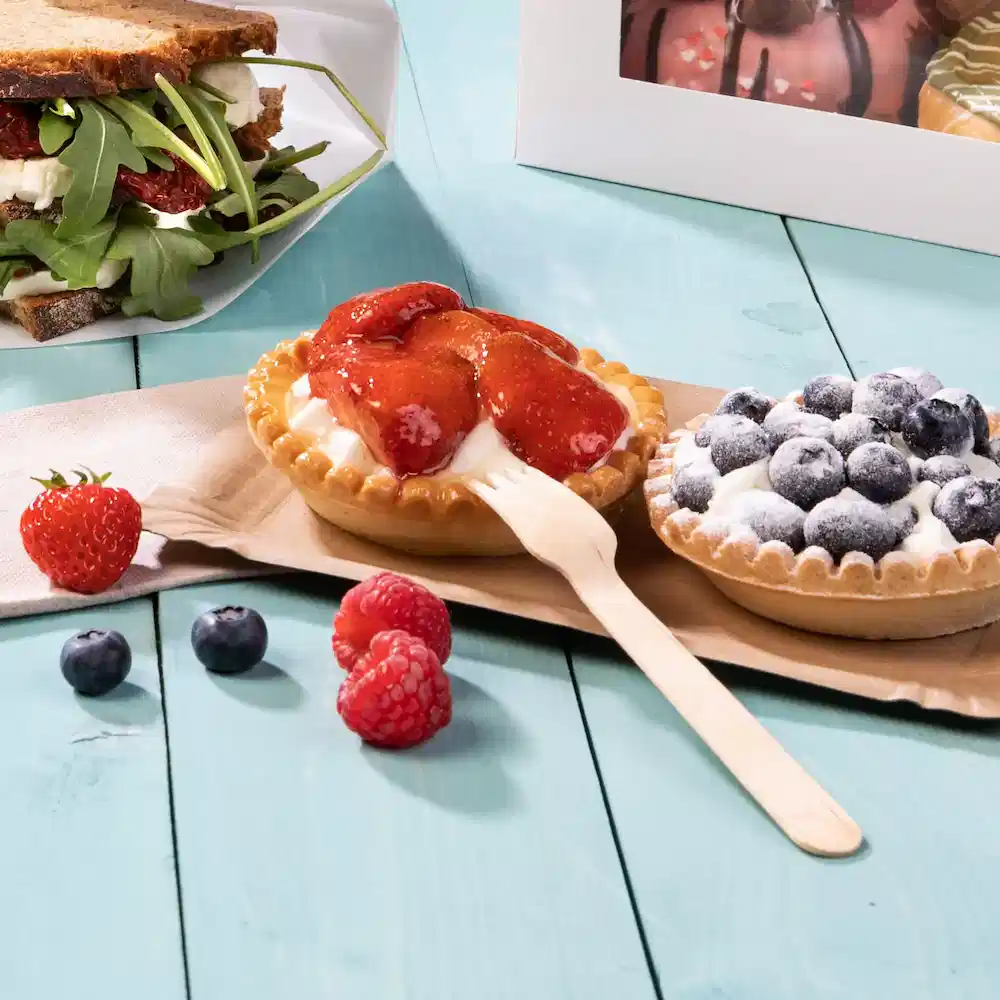 Two fruit tarts with strawberry and blueberry toppings, a wooden fork, fresh berries, and a sandwich in the background.