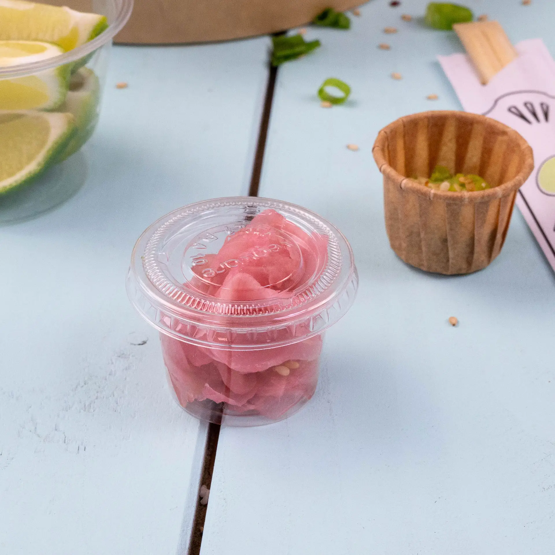 Clear plastic cup lid with pink pickled vegetables inside, placed on a light blue surface.