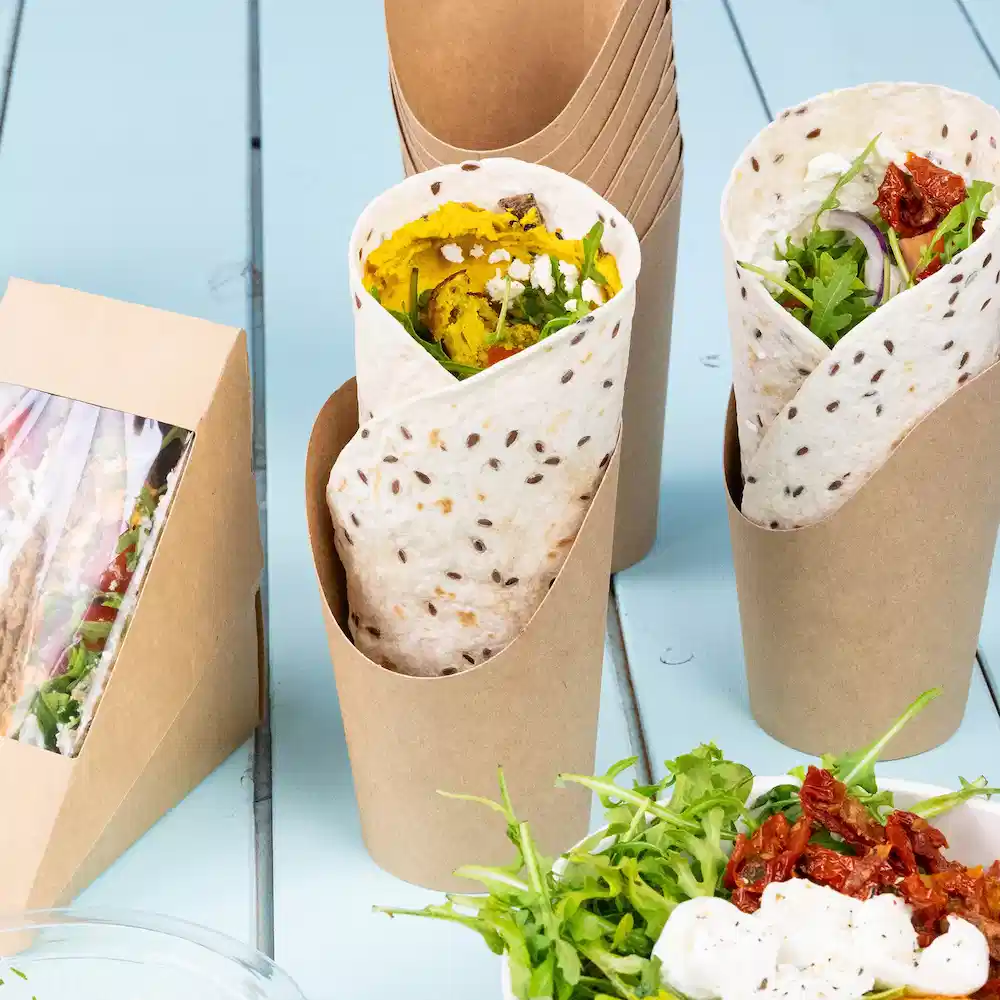 Two wraps in paper holders with ingredients visible, along with a side of salad and sun-dried tomatoes on a table.